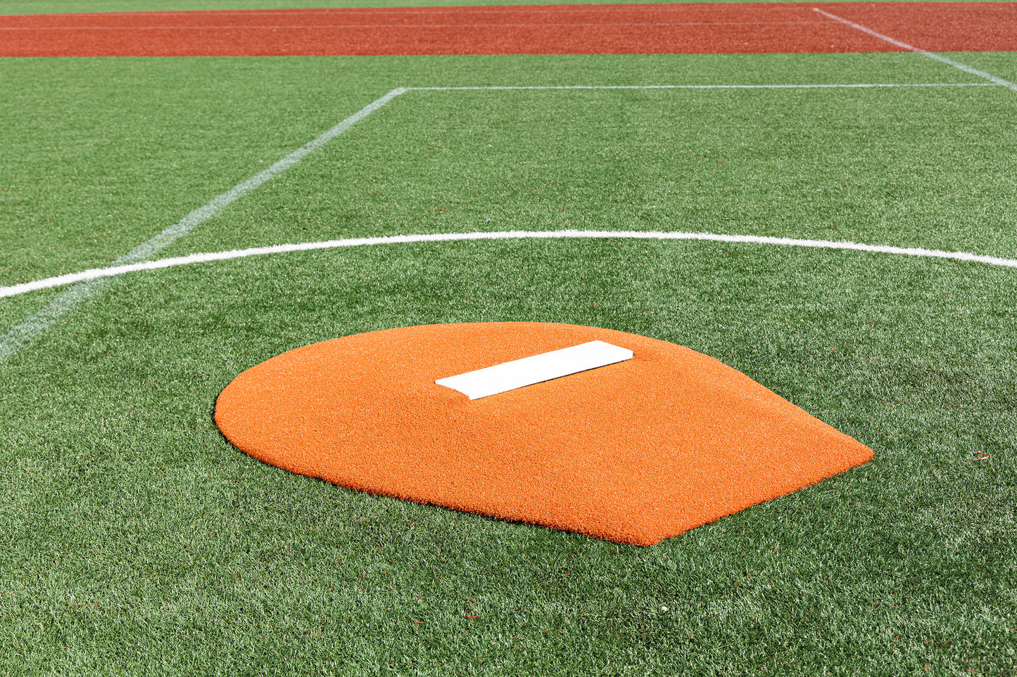 A close-up of the Portolite 6″ Stride Off Game Mound for ages 13 & under, featuring its bright orange design with a white rubber plate set on green turf—ideal for Little League pitchers. White boundary lines and red turf appear in the background.