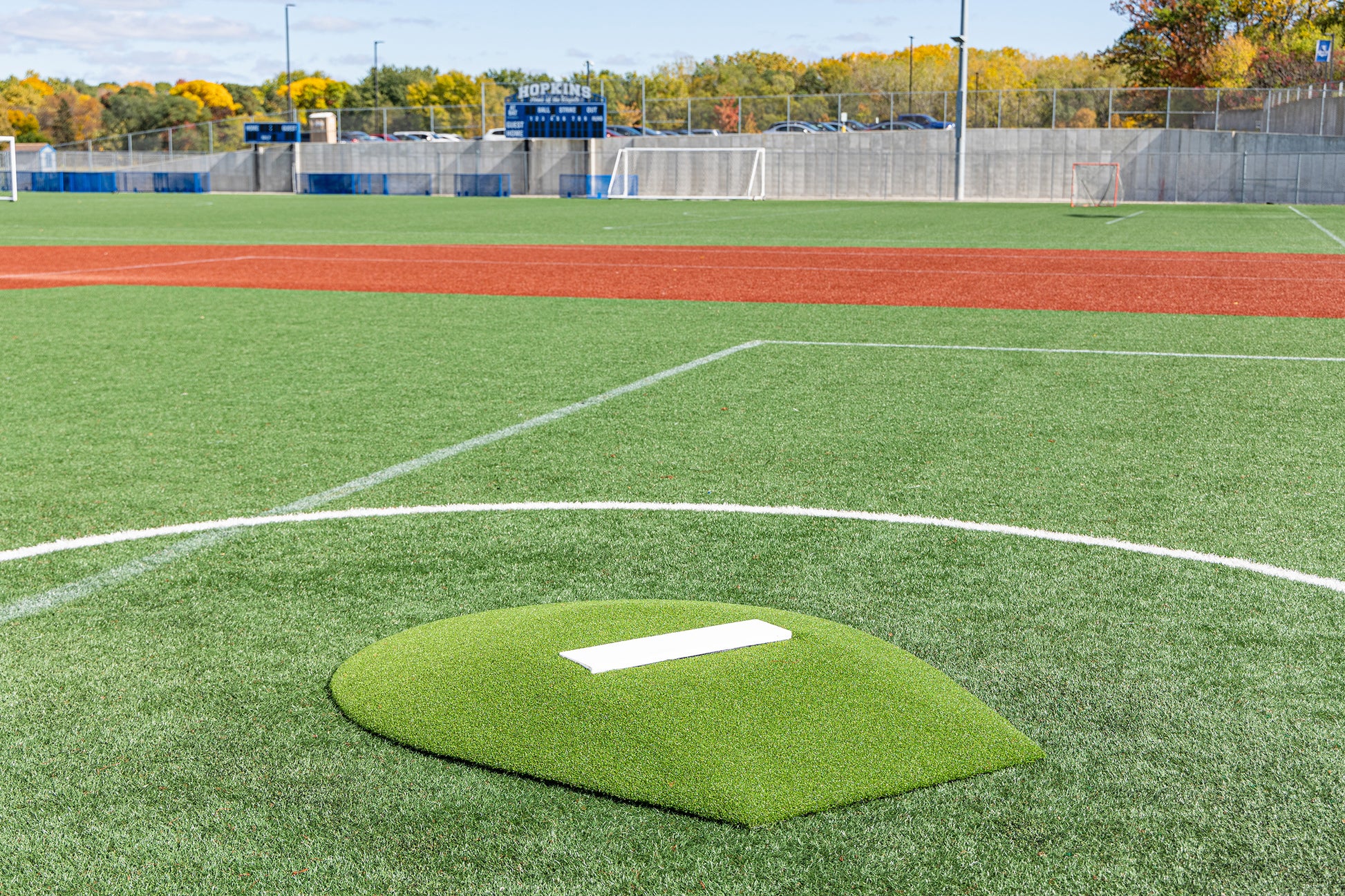 A synthetic turf baseball field featuring the Portolite 6″ Stride Off Game Mound—for ages 13 & under—in the foreground, perfect for youth pitchers, with a scoreboard, fence, and autumn trees in the background.
