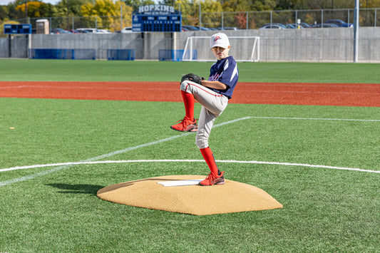 A young Little League baseball player in uniform stands mid-windup on the Portolite 6″ Stride Off Game Mound, for ages 13 & under, ready to pitch on a sunny day at a baseball field with a scoreboard and trees in the background.