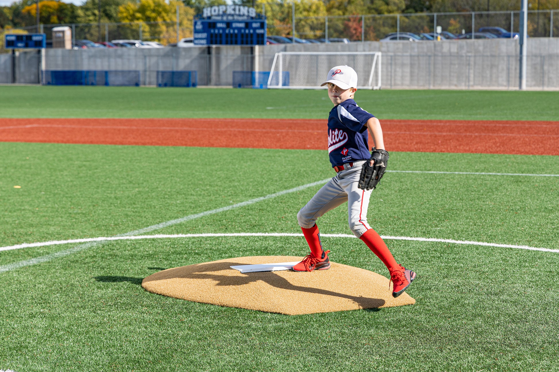 A young Little League baseball player in a blue jersey and red socks winds up on the Portolite 6″ Stride Off Game Mound (for ages 13 & under) at a sunny outdoor field, with a scoreboard and netted fence in the background.