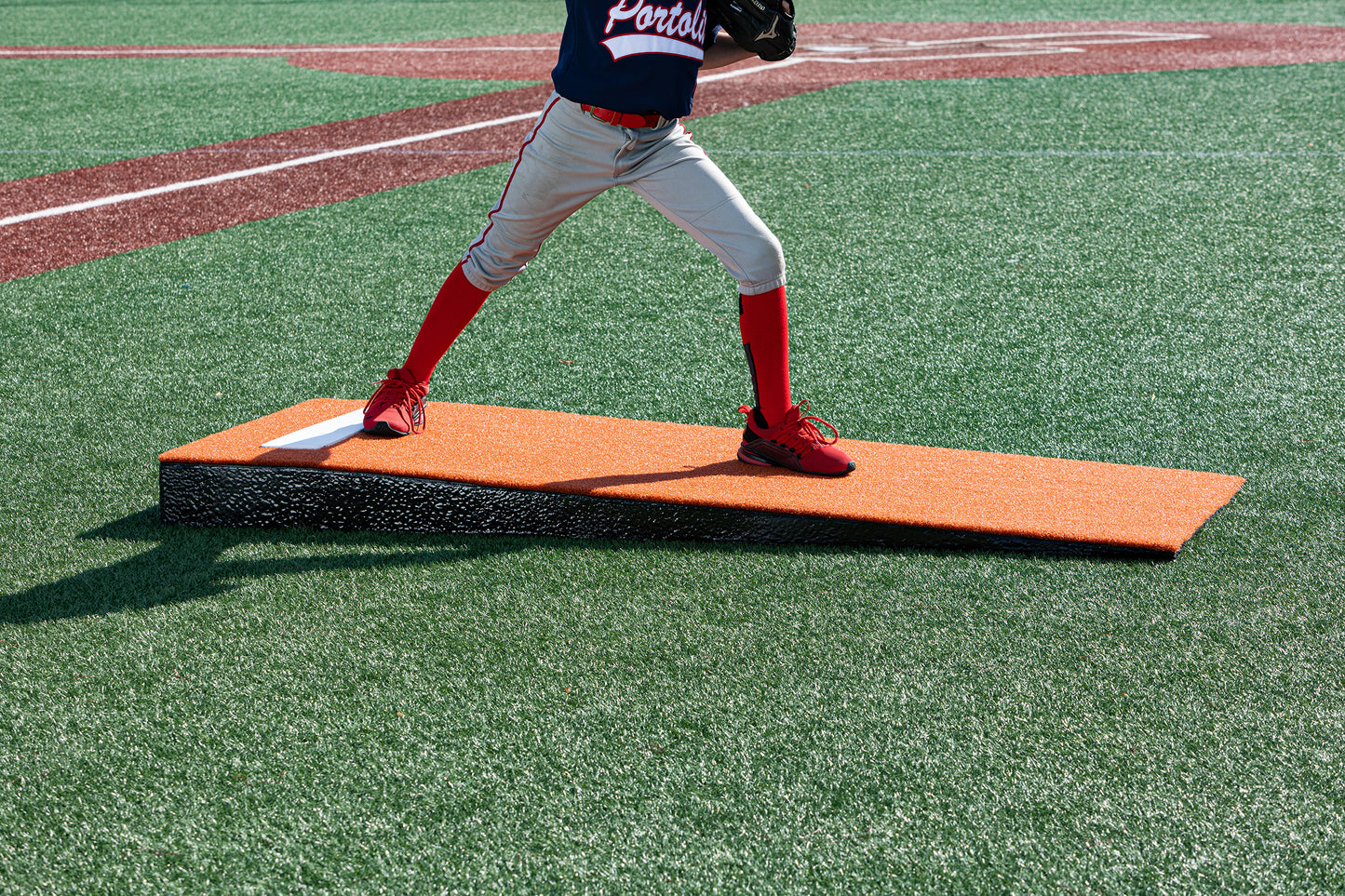 A young baseball player stands mid-pitch on the vivid orange Portolite Junior Practice Mound | Portable Pitching Mound for Under 12, their lower half visible as they train atop this youth baseball practice mound on an artificial turf field.