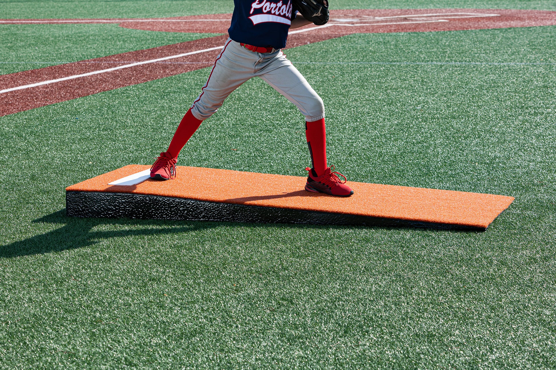 A young baseball player stands mid-pitch on the vivid orange Portolite Junior Practice Mound | Portable Pitching Mound for Under 12, their lower half visible as they train atop this youth baseball practice mound on an artificial turf field.