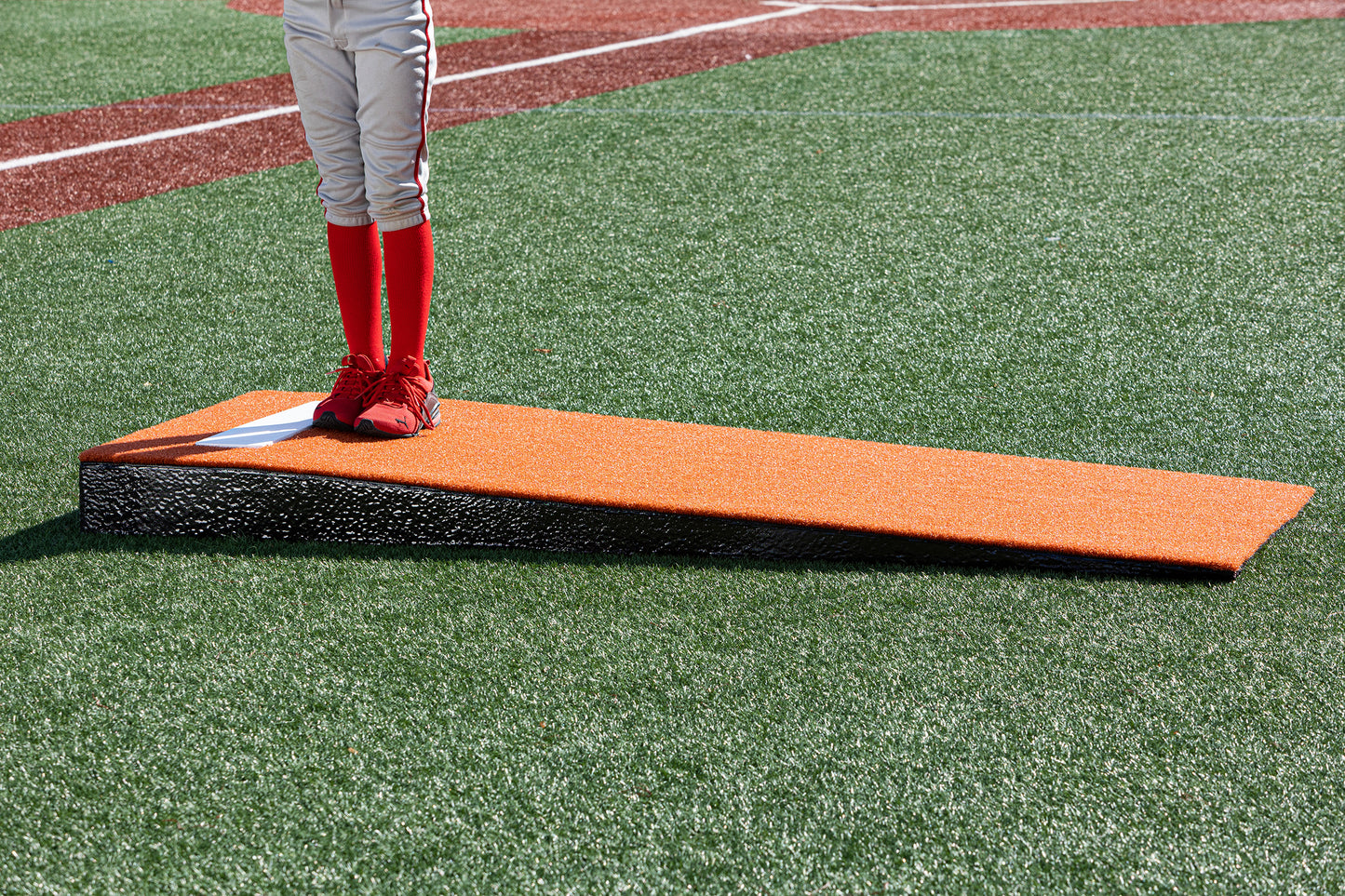 A child in red baseball pants and shoes stands on a Portolite Junior Practice Mound | Portable Pitching Mound for Under 12, set on an artificial turf baseball field.