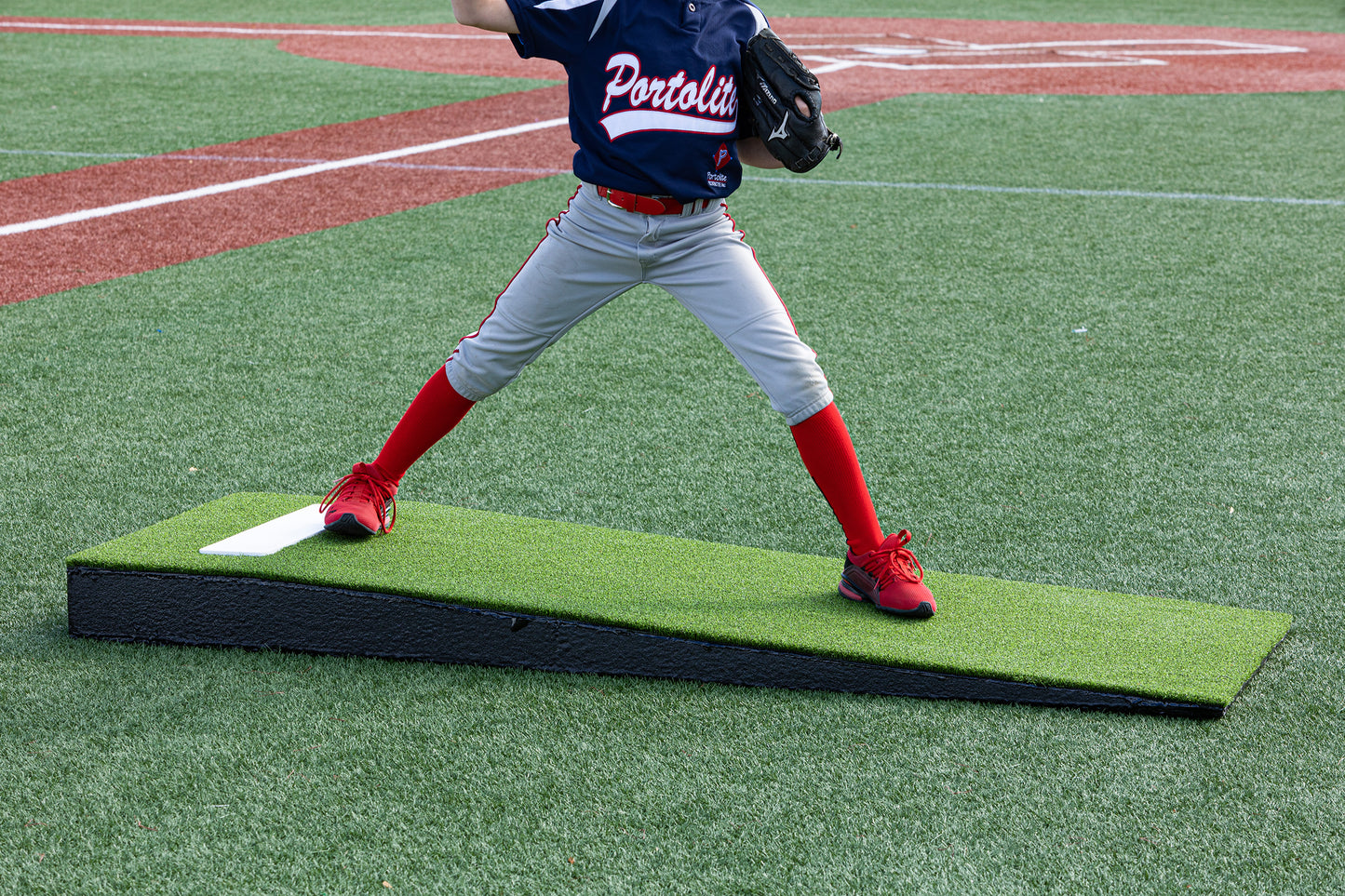 A young baseball player, visible from the shoulders down, stands with legs apart on a Portolite Junior Practice Mound | Portable Pitching Mound for Under 12, wearing a blue jersey, gray pants, and red socks and shoes, ready to pitch on a baseball field.