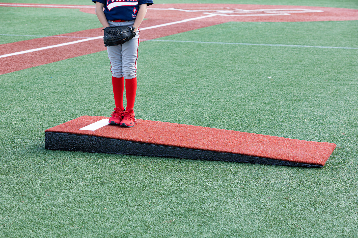 A young baseball player stands on a Portolite Junior Practice Mound portable pitching mound for under 12 on an artificial turf field. Only the player’s legs, dressed in gray pants and red socks, are visible; the rest of the player is out of frame.