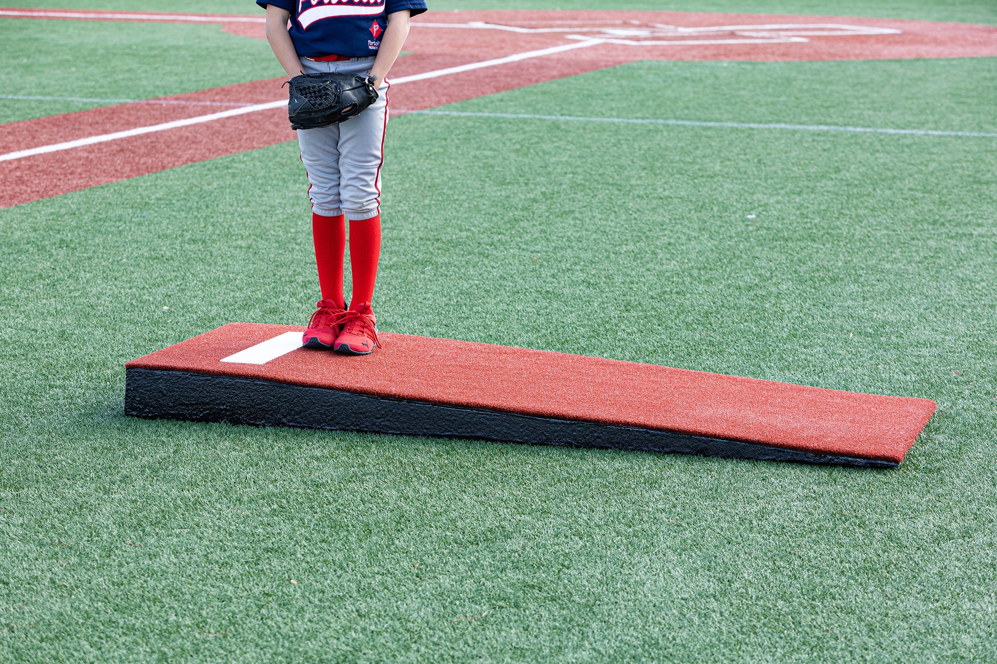A young baseball player stands on a Portolite Junior Practice Mound portable pitching mound for under 12 on an artificial turf field. Only the player’s legs, dressed in gray pants and red socks, are visible; the rest of the player is out of frame.