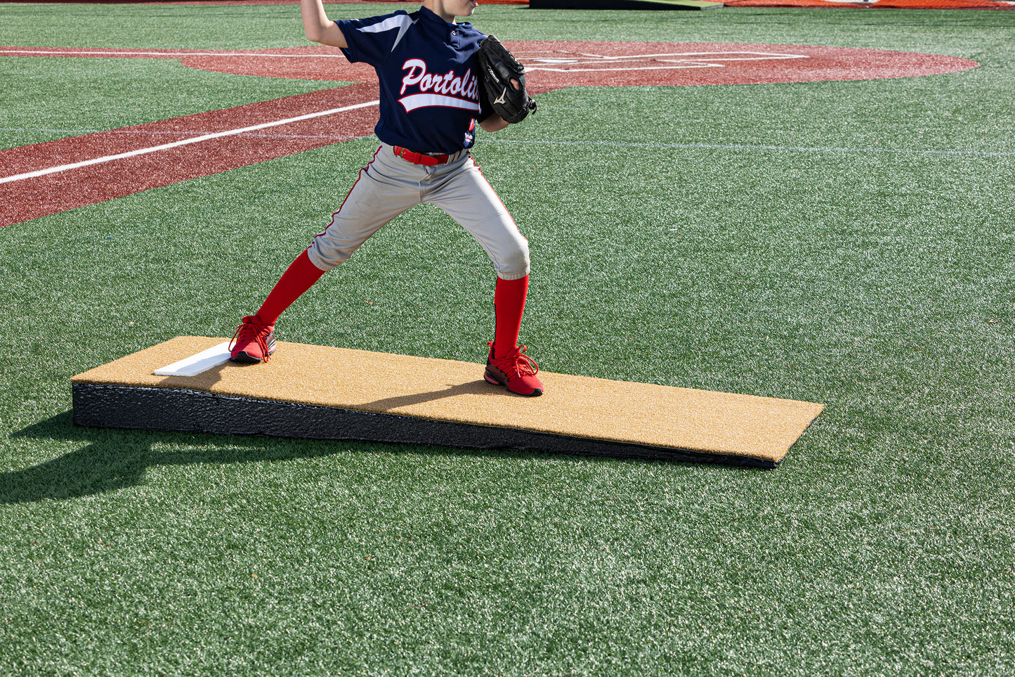 A young baseball pitcher, visible from the waist down in red socks and a navy jersey, throws a pitch on a Portolite Junior Practice Mound | Portable Pitching Mound for Under 12 on a synthetic turf field. The scene highlights how this portable pitching mound is ideal for youth baseball players.