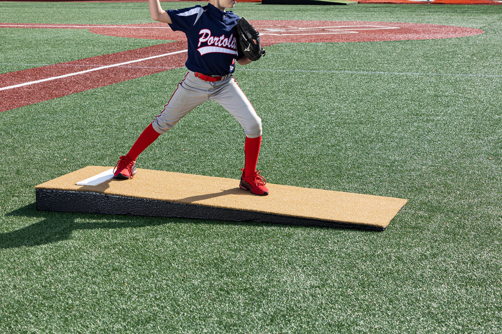 A young baseball pitcher, visible from the waist down in red socks and a navy jersey, throws a pitch on a Portolite Junior Practice Mound | Portable Pitching Mound for Under 12 on a synthetic turf field. The scene highlights how this portable pitching mound is ideal for youth baseball players.