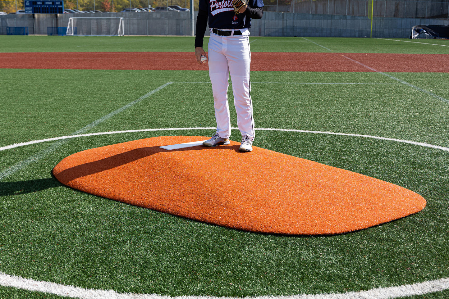 A baseball player stands on a Portolite 8″ One-Piece Game Mound | Ages 13-15, its orange surface contrasting with the synthetic green field—an ideal setup for multi-age tournaments or Babe Ruth level games. The player’s upper body is out of frame. White lines mark the pitching area, and bases are visible in the background.