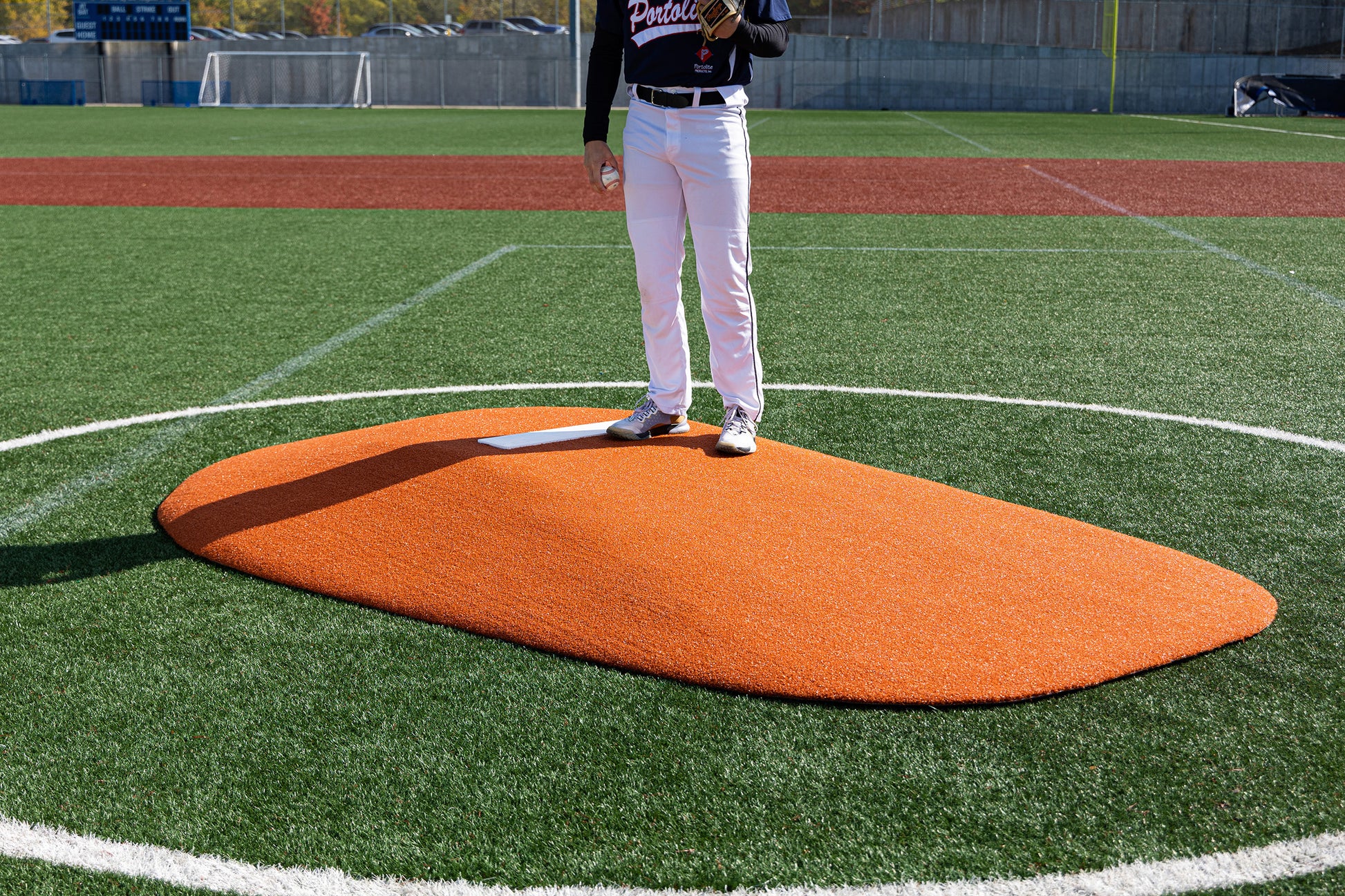 A baseball player stands on a Portolite 8″ One-Piece Game Mound | Ages 13-15, its orange surface contrasting with the synthetic green field—an ideal setup for multi-age tournaments or Babe Ruth level games. The player’s upper body is out of frame. White lines mark the pitching area, and bases are visible in the background.