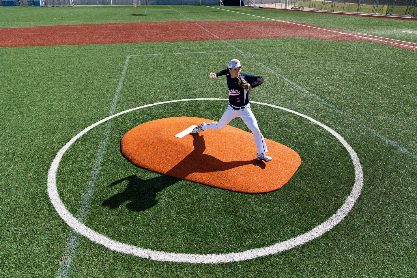 A baseball pitcher in a black jersey and white pants winds up on the Portolite 8″ One-Piece Game Mound for Ages 13-15, positioned within a white pitchers circle on an artificial turf field in daylight—an ideal setup for multi-age tournaments or Babe Ruth-level games.