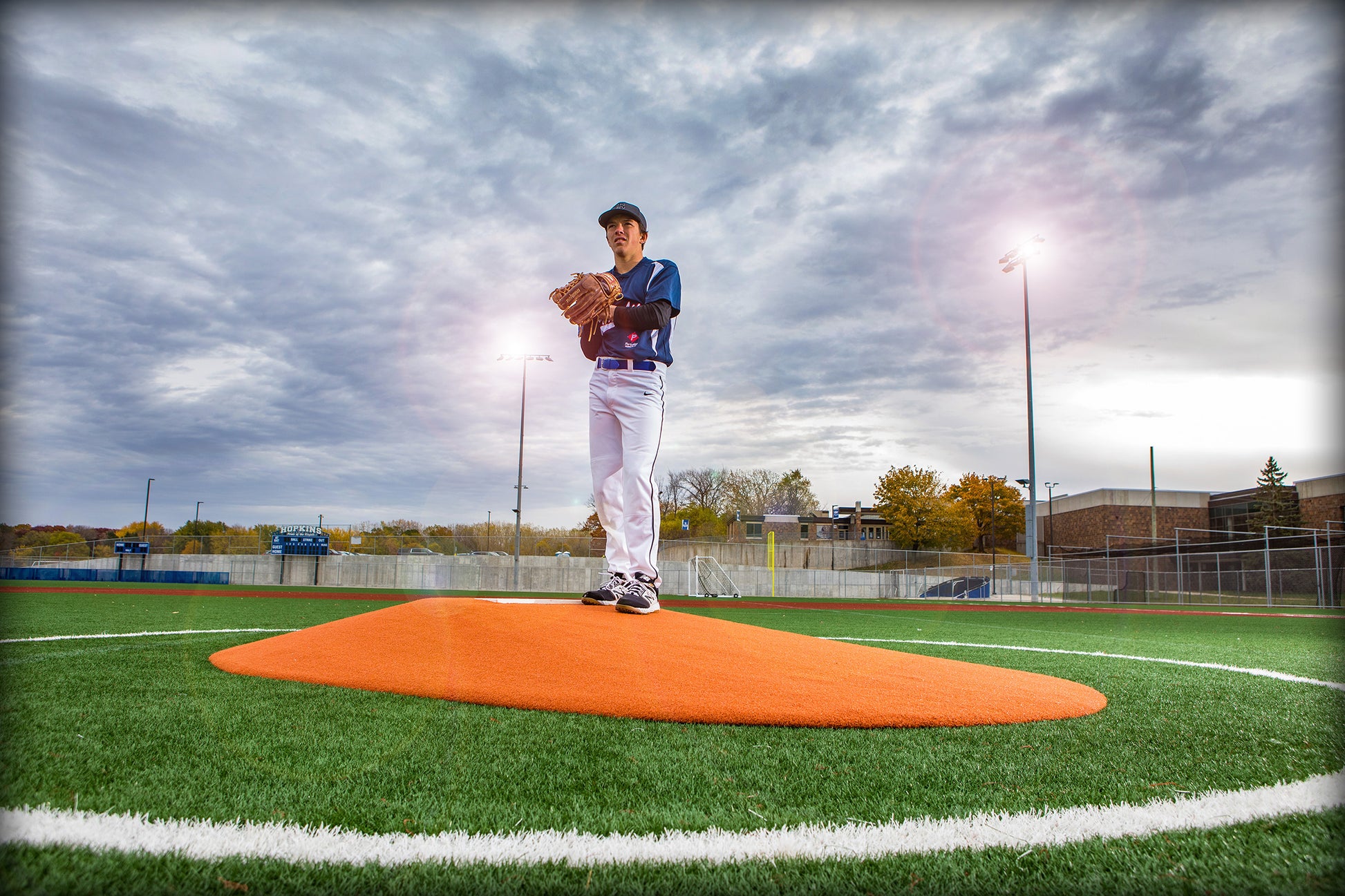 A young baseball player stands on a Portolite 8″ One-Piece Game Mound (for ages 13-15), preparing to pitch beneath a cloudy sky on an outdoor field, with stadium lights and buildings in the background—the perfect setting for Babe Ruth level and multi-age tournaments.