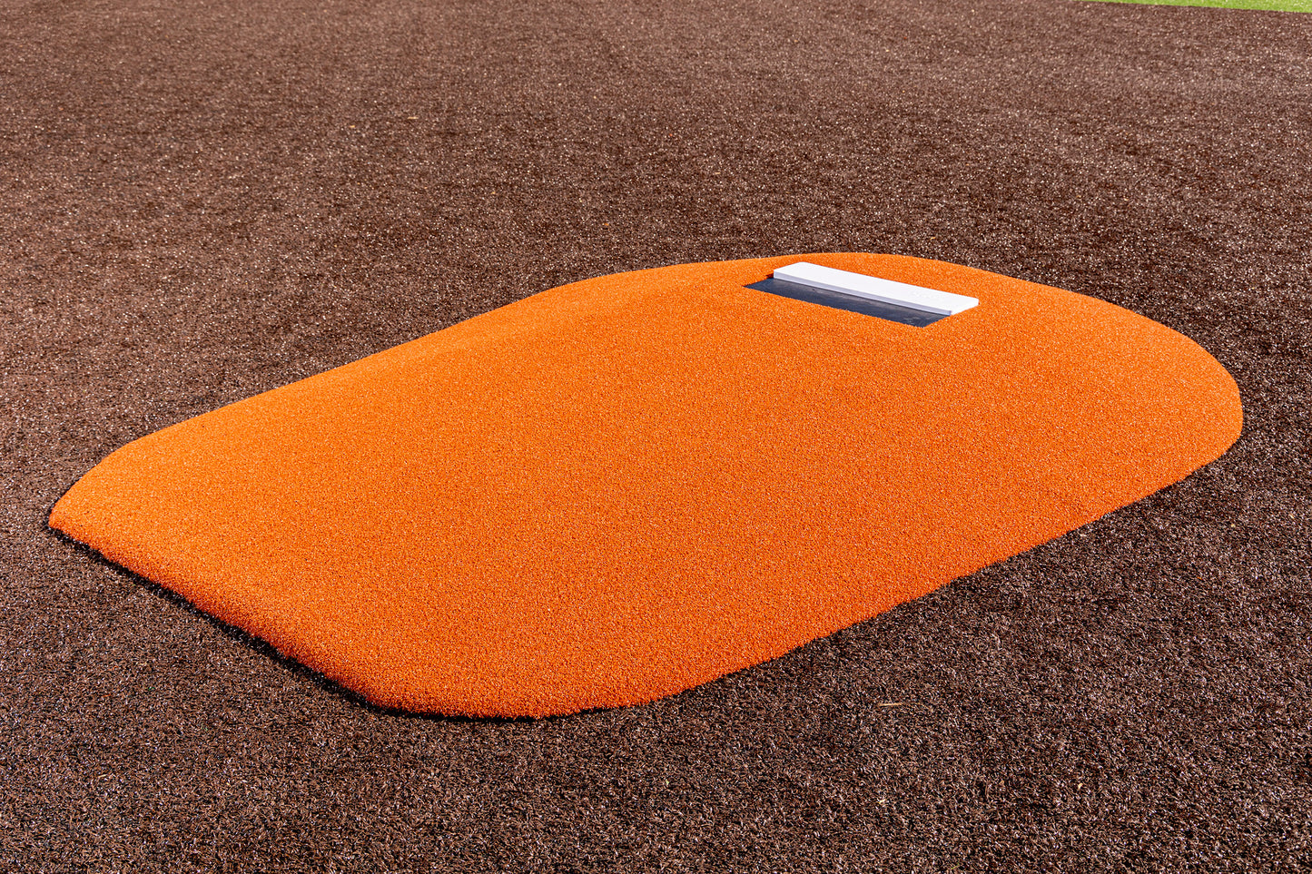 A bright orange Portolite 8″ One-Piece Game Mound for ages 13-15, featuring a white rubber plate, sits on a brown artificial turf field—perfect for Babe Ruth level play and multi-age tournaments.