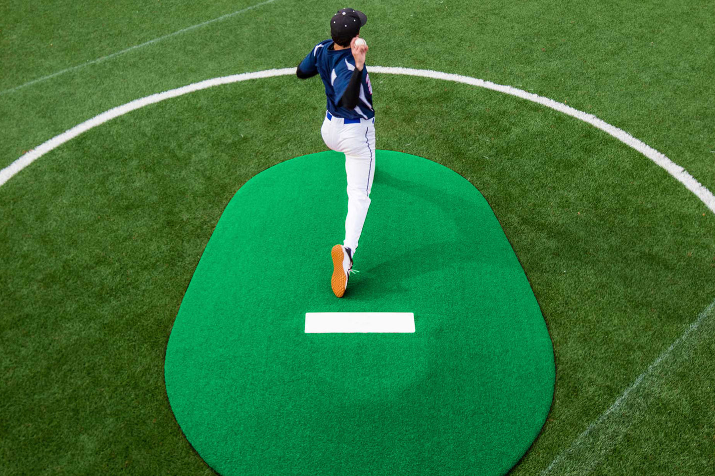 A baseball player in uniform pitches from a green Portolite 8″ One-Piece Game Mound (for ages 13-15) on an artificial turf field, viewed from above—a setup ideal for multi-age tournaments and perfect for Babe Ruth level play.