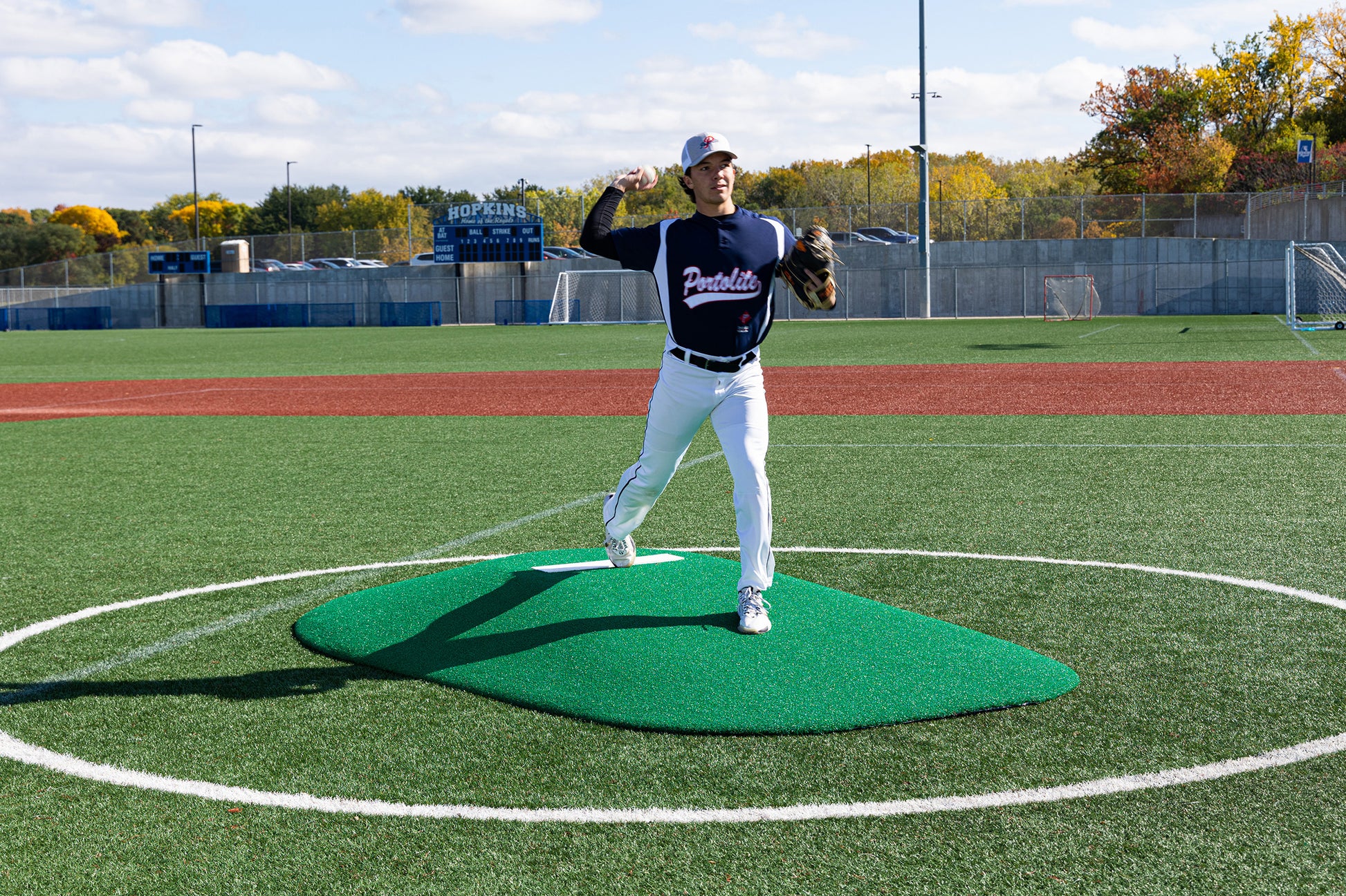 A baseball player in a dark jersey and white pants stands atop the Portolite 8″ One-Piece Game Mound | Ages 13-15, ready to pitch on a bright, sunny day at an outdoor stadium—perfect for competitive play or multi-age tournaments.