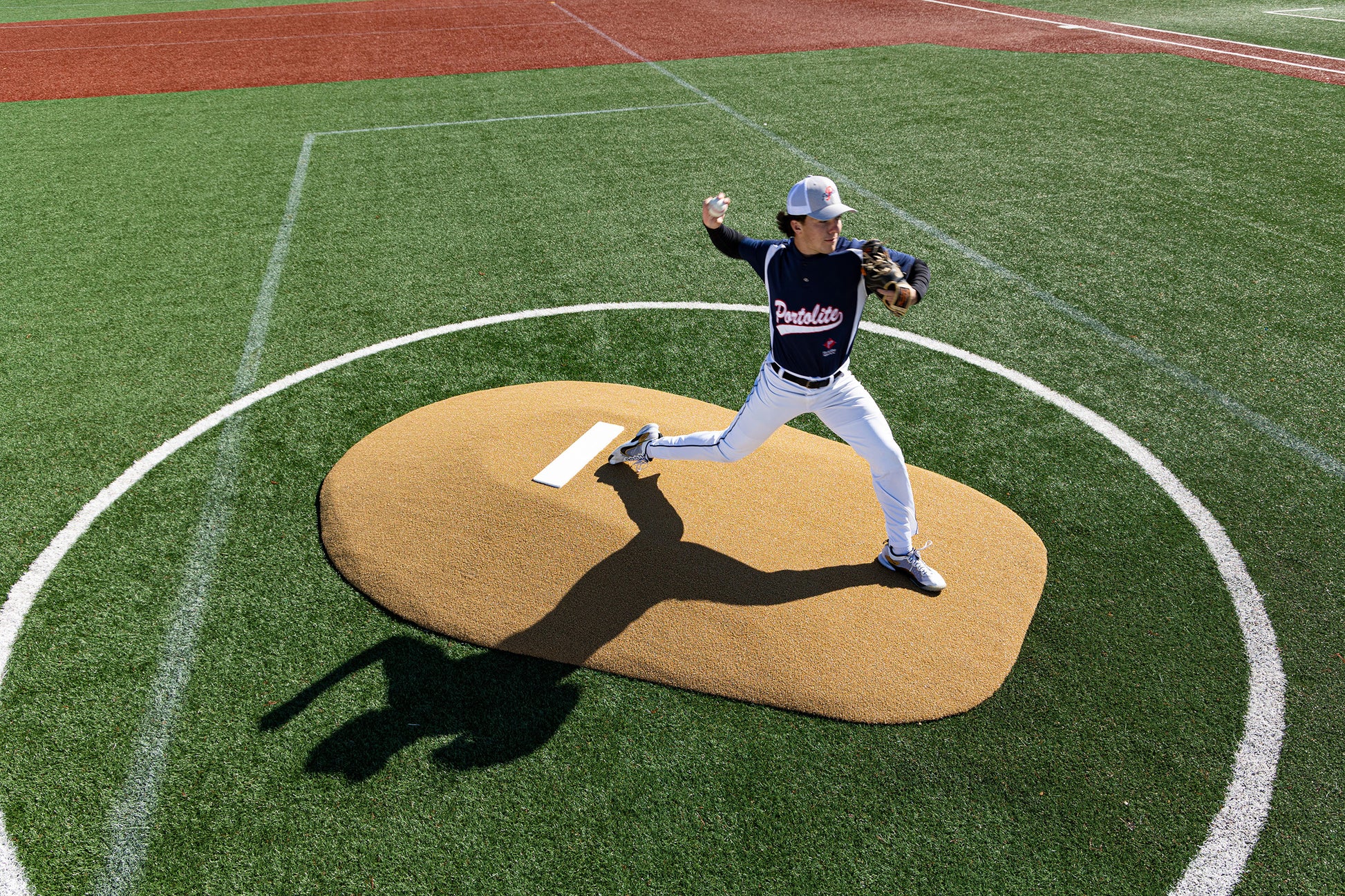 A baseball pitcher in a navy Panthers jersey winds up to throw a pitch from the Portolite 8″ One-Piece Game Mound (for ages 13-15) on a Babe Ruth-level artificial turf field, casting a shadow as he competes in one of the multi-age tournaments.