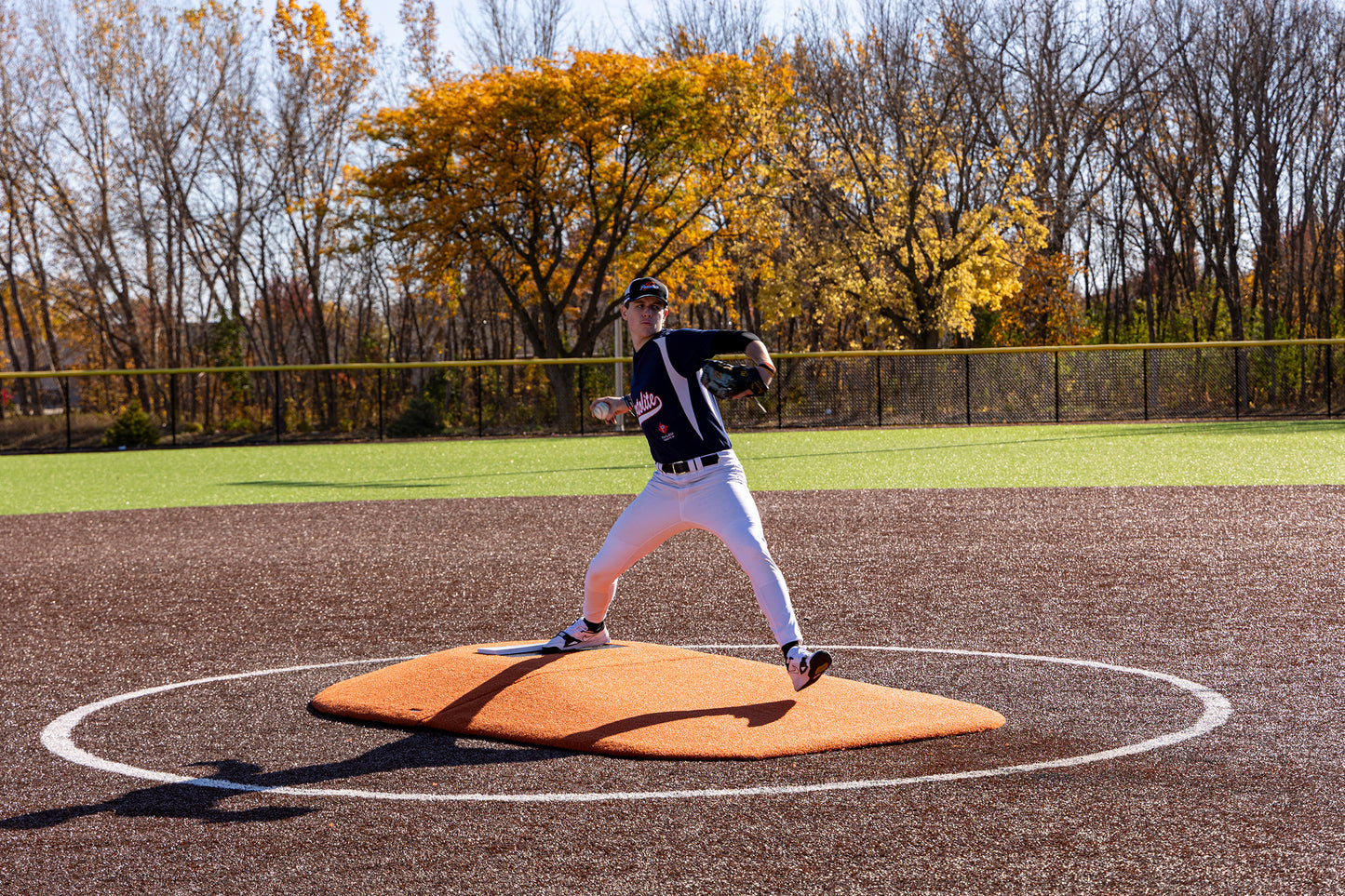 A baseball pitcher in uniform is mid-throw on the Portolite 8″ Two-Piece Game Mound (for ages 13–15), with autumn trees and a fence in the background under a clear sky.
