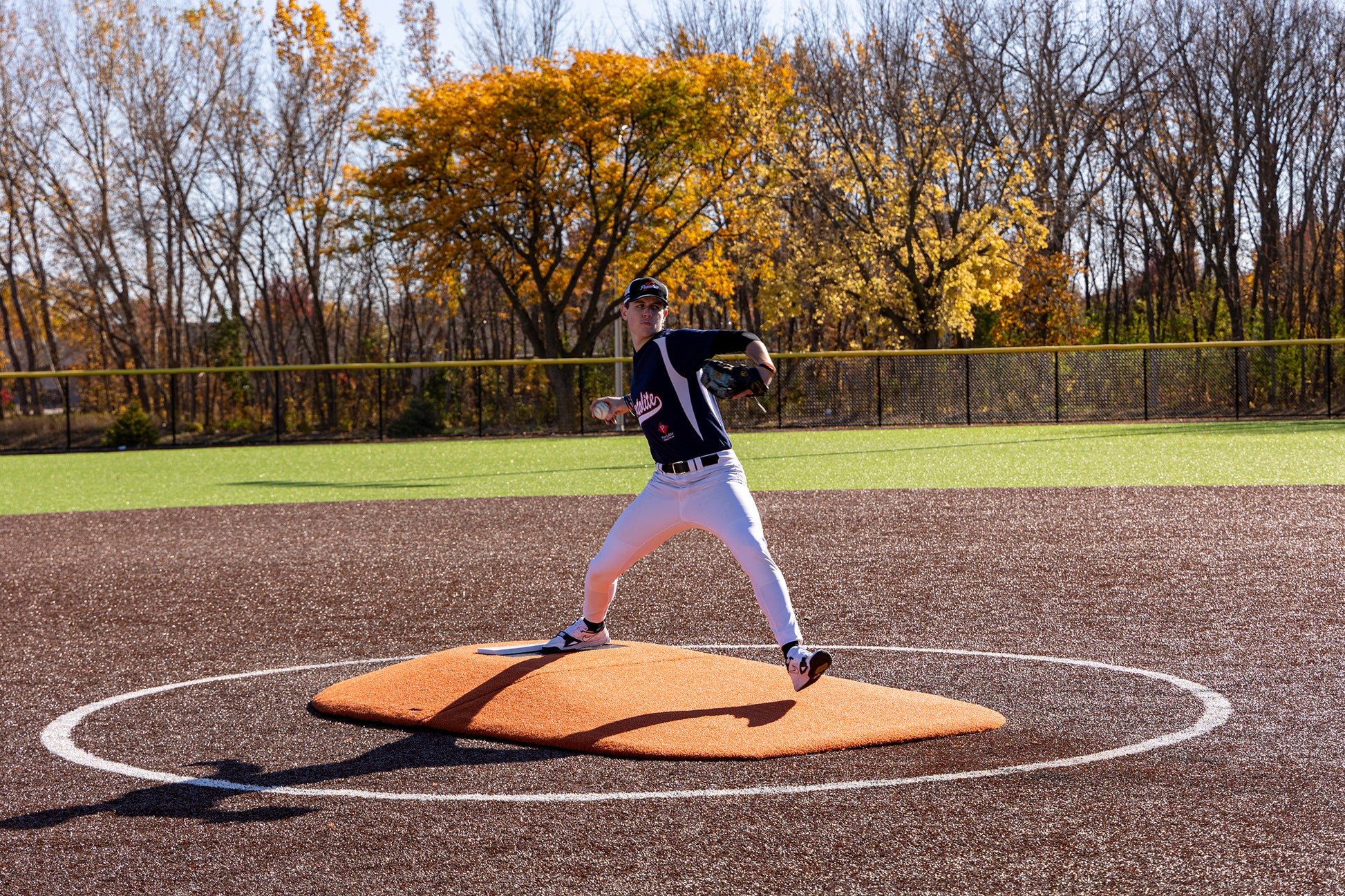 A baseball pitcher in uniform is mid-throw on the Portolite 8″ Two-Piece Game Mound (for ages 13–15), with autumn trees and a fence in the background under a clear sky.