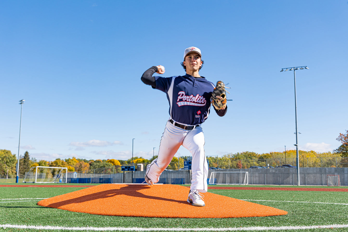 A young baseball pitcher in a navy Portolite uniform winds up to throw from the Portolite 8″ Two-Piece Game Mound, designed for ages 13-15, on a turf field under a clear blue sky.