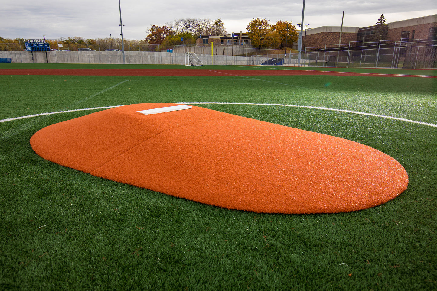 The Portolite 8″ Two-Piece Game Mound for ages 13-15, featuring a vibrant orange color and white rubber plate, sits on green artificial turf under a cloudy sky—ideal for Babe Ruth level games and transitional mound training.