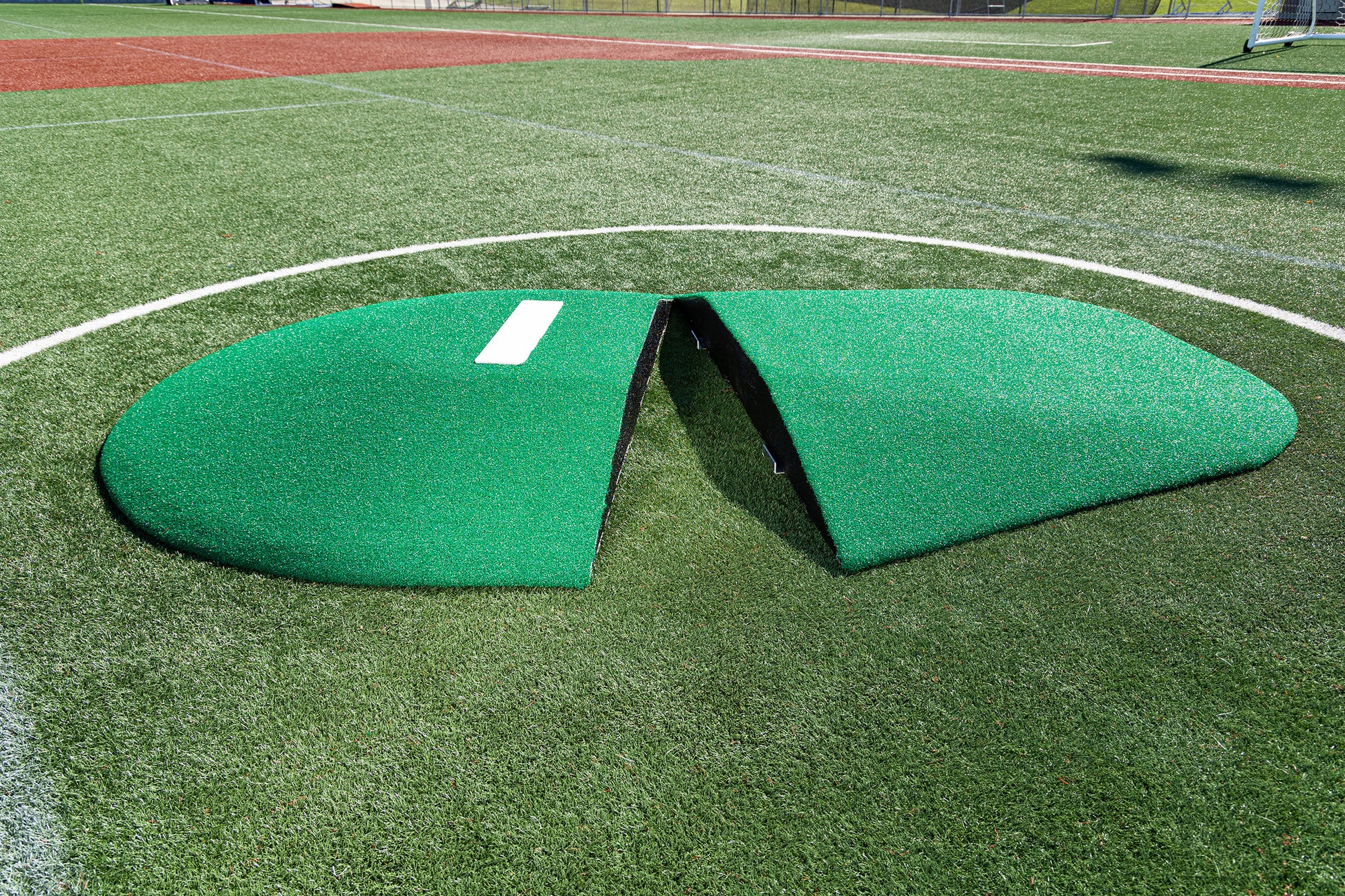 The Portolite 8″ Two-Piece Game Mound for ages 13-15 sits on an artificial turf baseball field inside the pitching circle. Split in half for easy portability, this green mound is ideal for Babe Ruth level play, offering flexibility and convenience for youth leagues.