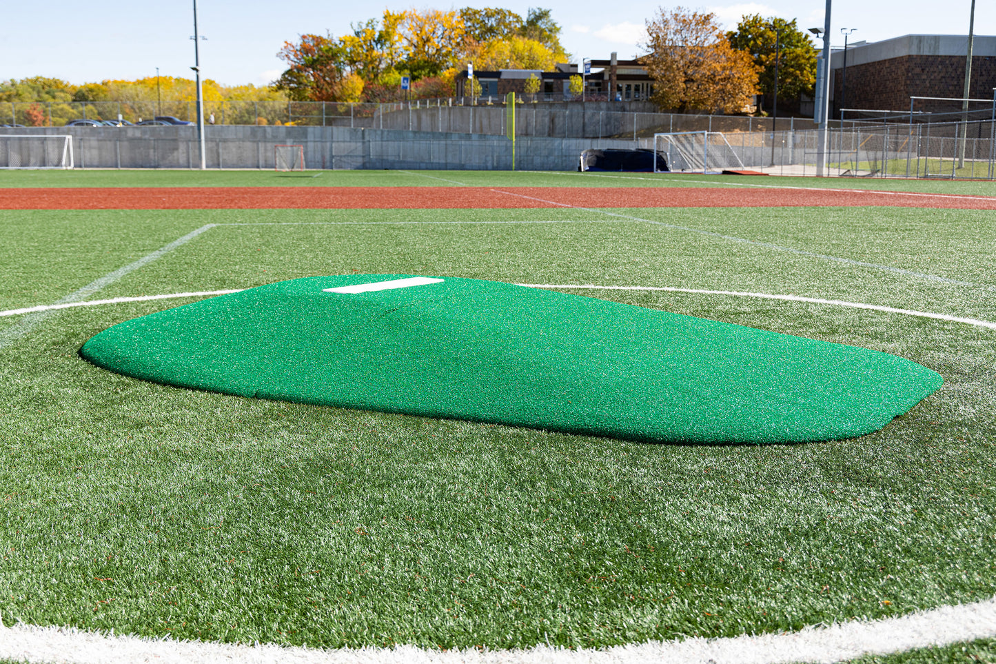 A close-up of the Portolite 8″ Two-Piece Game Mound, designed for ages 13-15, set on an artificial turf baseball field and encircled by a white line, with trees and a building in the background—ideal for Babe Ruth level play.