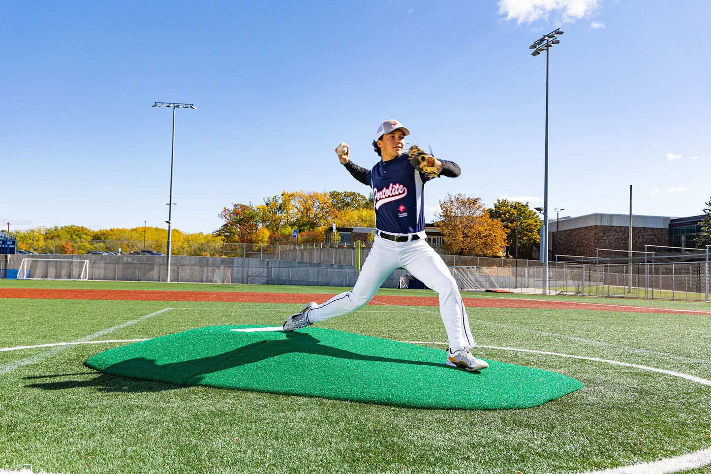 A baseball pitcher in uniform winds up to throw from the Portolite 8″ Two-Piece Game Mound (for ages 13-15) on an outdoor field under a clear blue sky, with autumn trees in the background—perfect for Babe Ruth-level games.