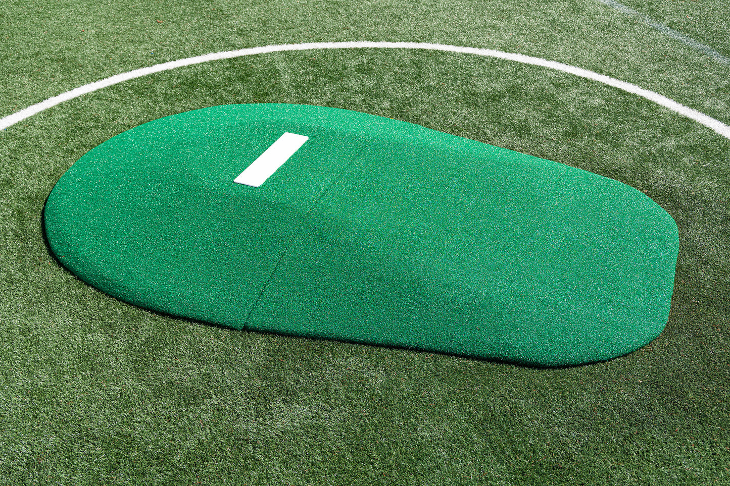 A Portolite 8″ Two-Piece Game Mound for ages 13-15, colored green with a white pitching rubber, sits on a synthetic turf baseball field with part of the pitcher’s circle visible. This portable mound is ideal for Babe Ruth level play and serves as an excellent transitional mound for developing athletes.