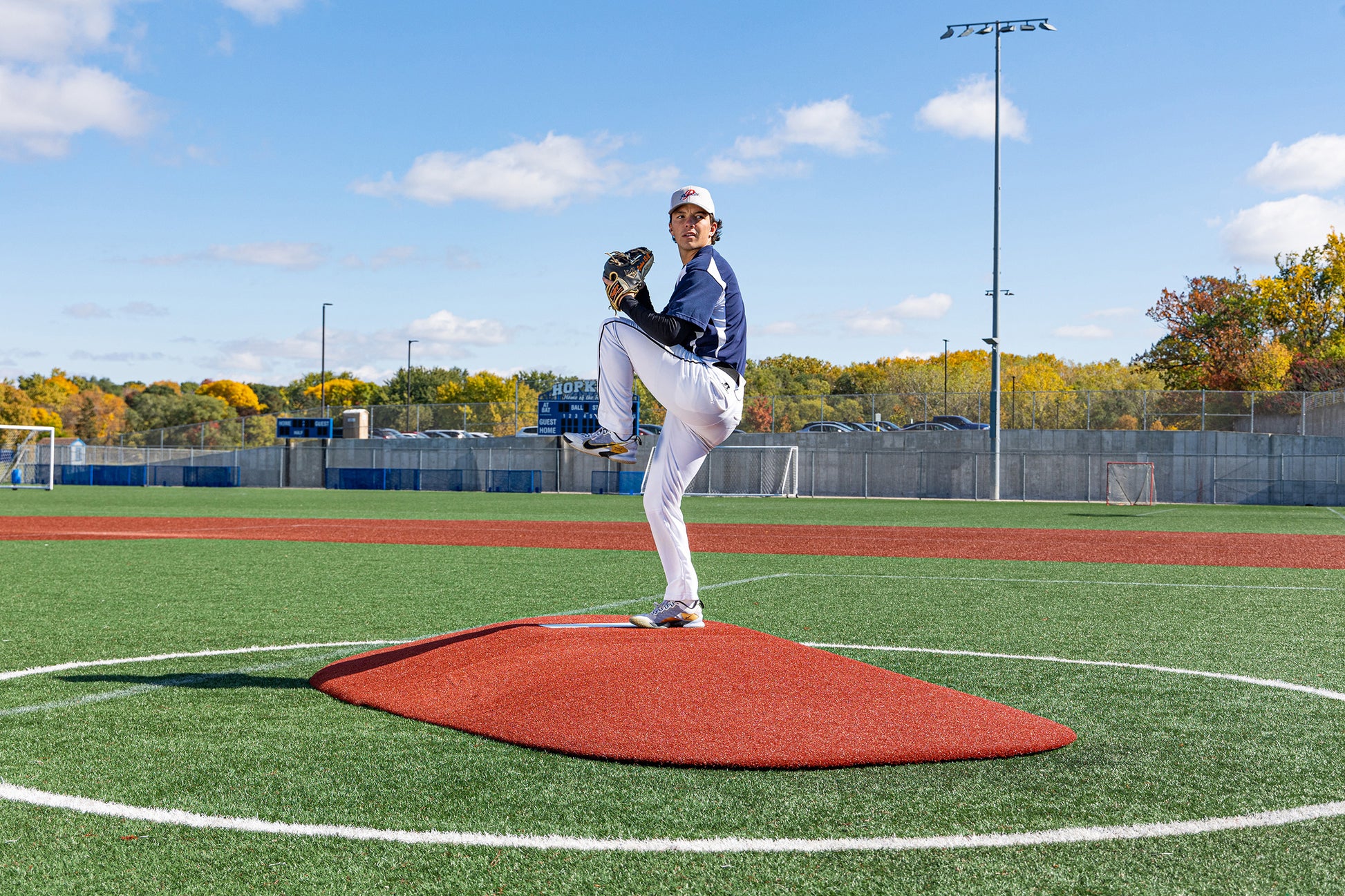 A baseball pitcher stands mid-windup on the Portolite 8″ Two-Piece Game Mound for ages 13-15, getting ready to deliver a pitch on an outdoor field with blue sky and autumn trees in the background, demonstrating Babe Ruth-level skills.