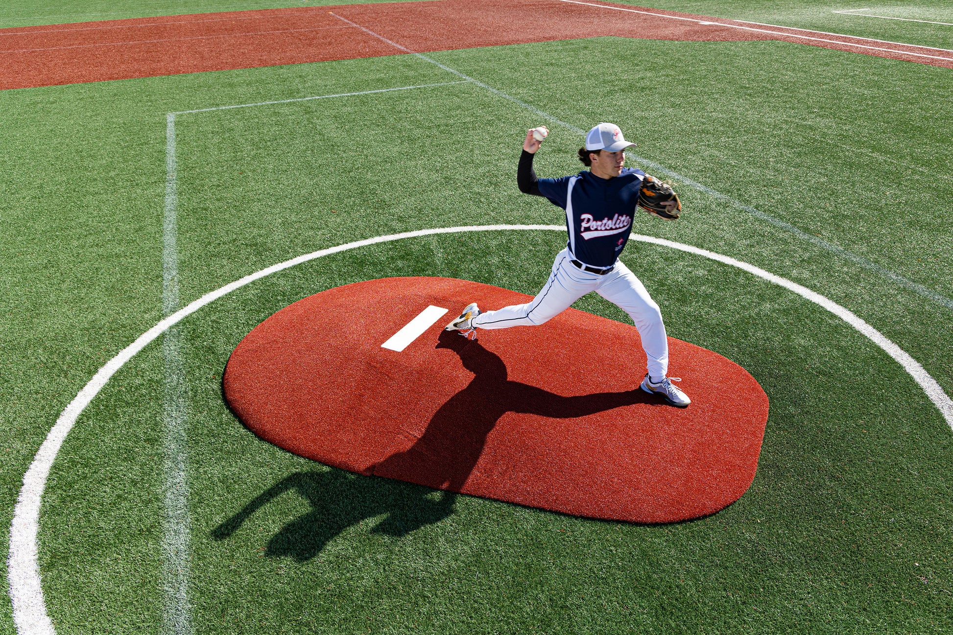 A baseball pitcher in a navy uniform, wearing a white cap, prepares to throw with an outstretched arm while standing on the Portolite 8″ Two-Piece Game Mound at a baseball field—ideal for transitional use at the Babe Ruth level for ages 13–15.