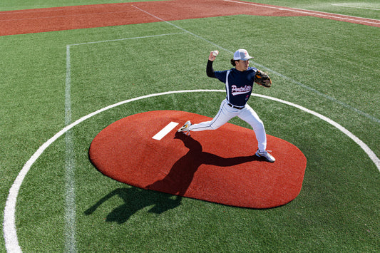 A baseball pitcher in a navy uniform, wearing a white cap, prepares to throw with an outstretched arm while standing on the Portolite 8″ Two-Piece Game Mound at a baseball field—ideal for transitional use at the Babe Ruth level for ages 13–15.