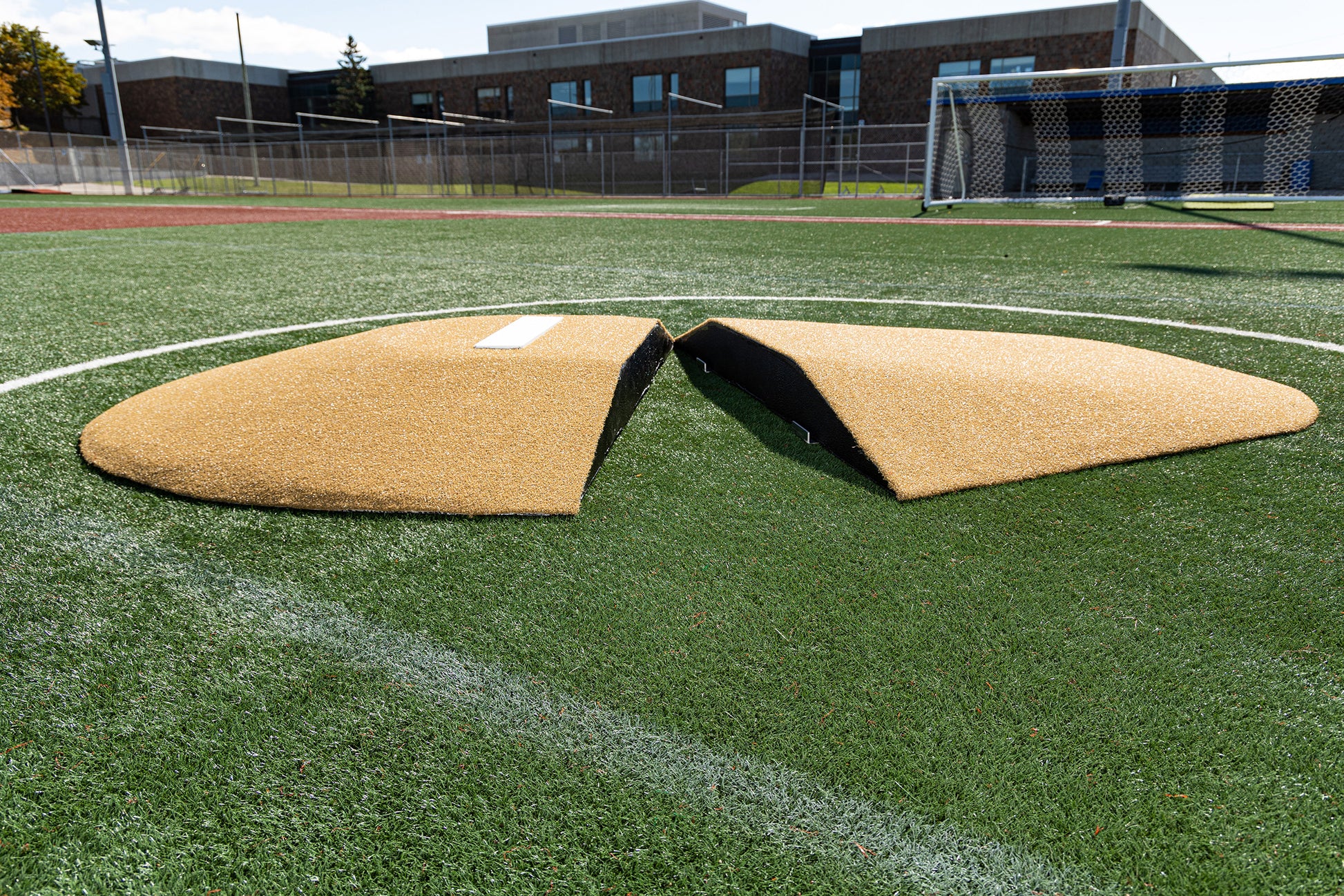 Two curved, tan-colored sections of the Portolite 8″ Two-Piece Game Mound for ages 13-15, featuring a white stripe, are positioned apart on a green sports field within a circular line near a goal post. This transitional mound is designed specifically for Babe Ruth level play, with a school building and fence visible in the background.