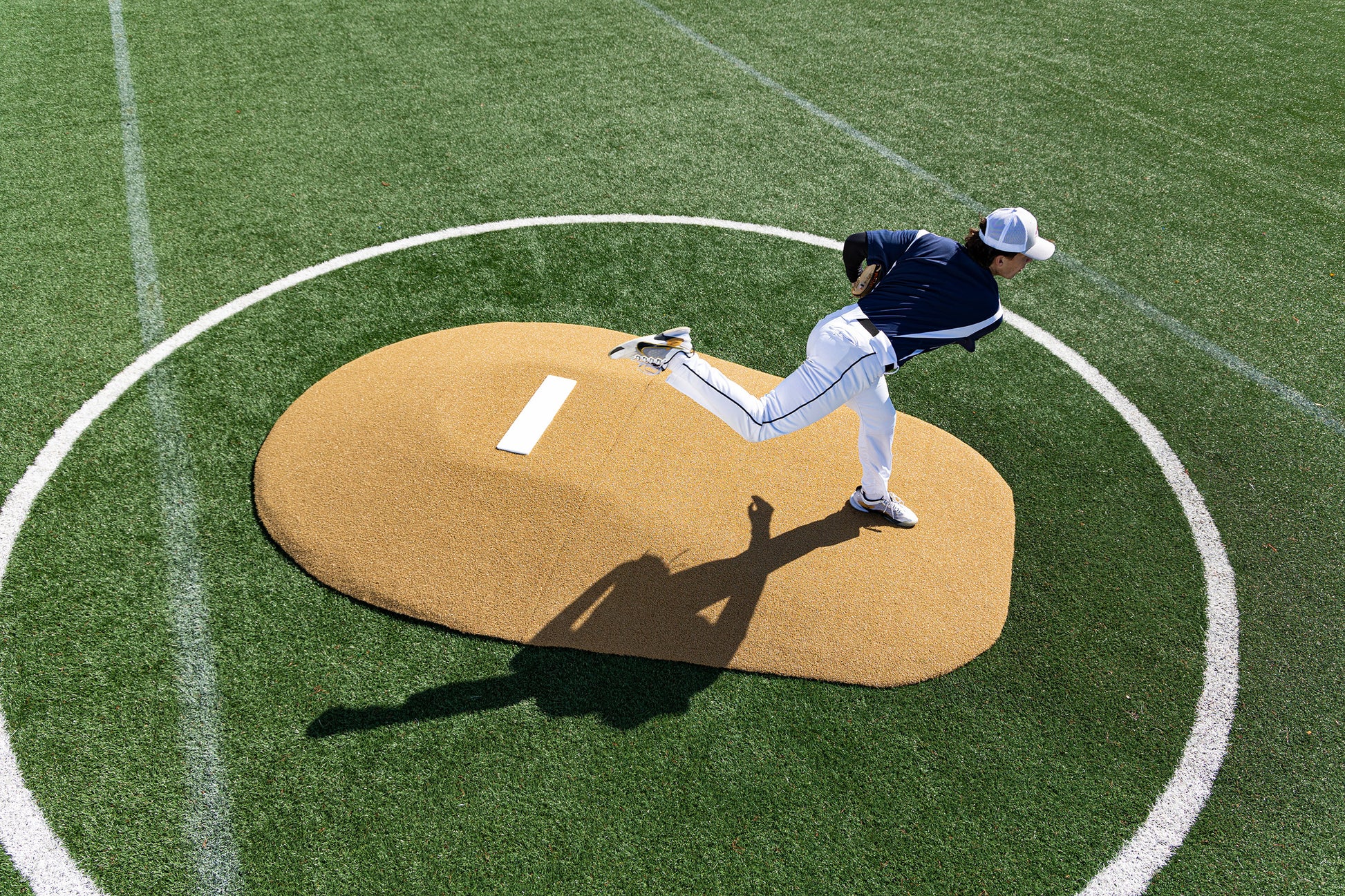 A baseball pitcher in a navy jersey and white pants winds up to throw from the Portolite 8″ Two-Piece Game Mound (for ages 13-15), viewed from above on a green turf field, ready for action at the Babe Ruth level.