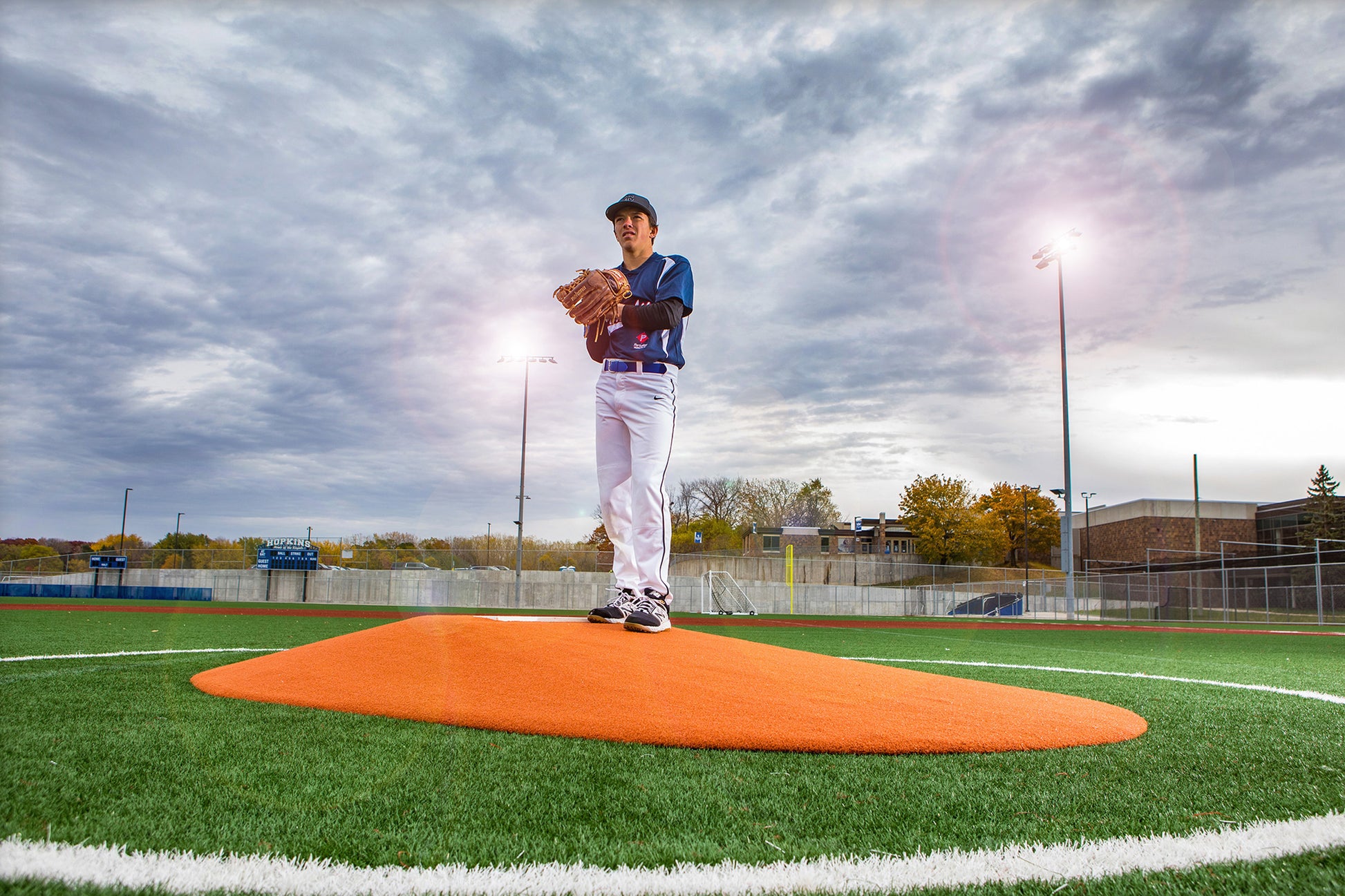 A young baseball player stands on a Portolite 10″ One-Piece Game Mound, a heavy-duty regulation pitching mound in bright orange, preparing to pitch on an outdoor field beneath a cloudy sky with stadium lights glowing in the background.