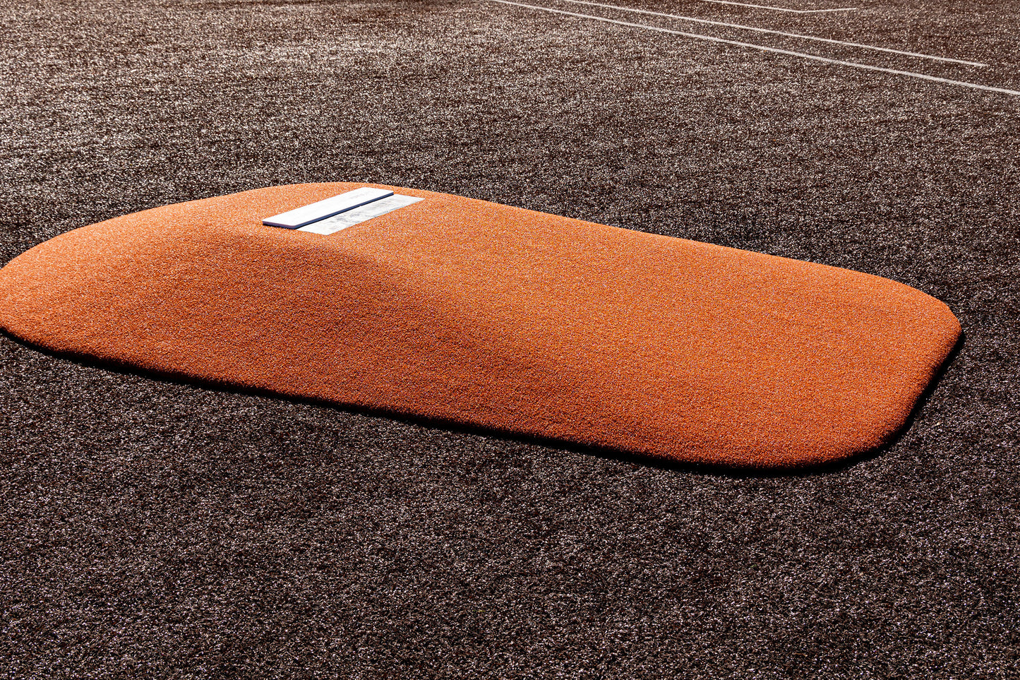 The Portolite 10″ One-Piece Game Mound, a heavy-duty regulation pitching mound featuring an orange design with a white rubber plate, is positioned on a brown artificial turf baseball field; visible white foul lines in the background make it ideal for high school baseball games.