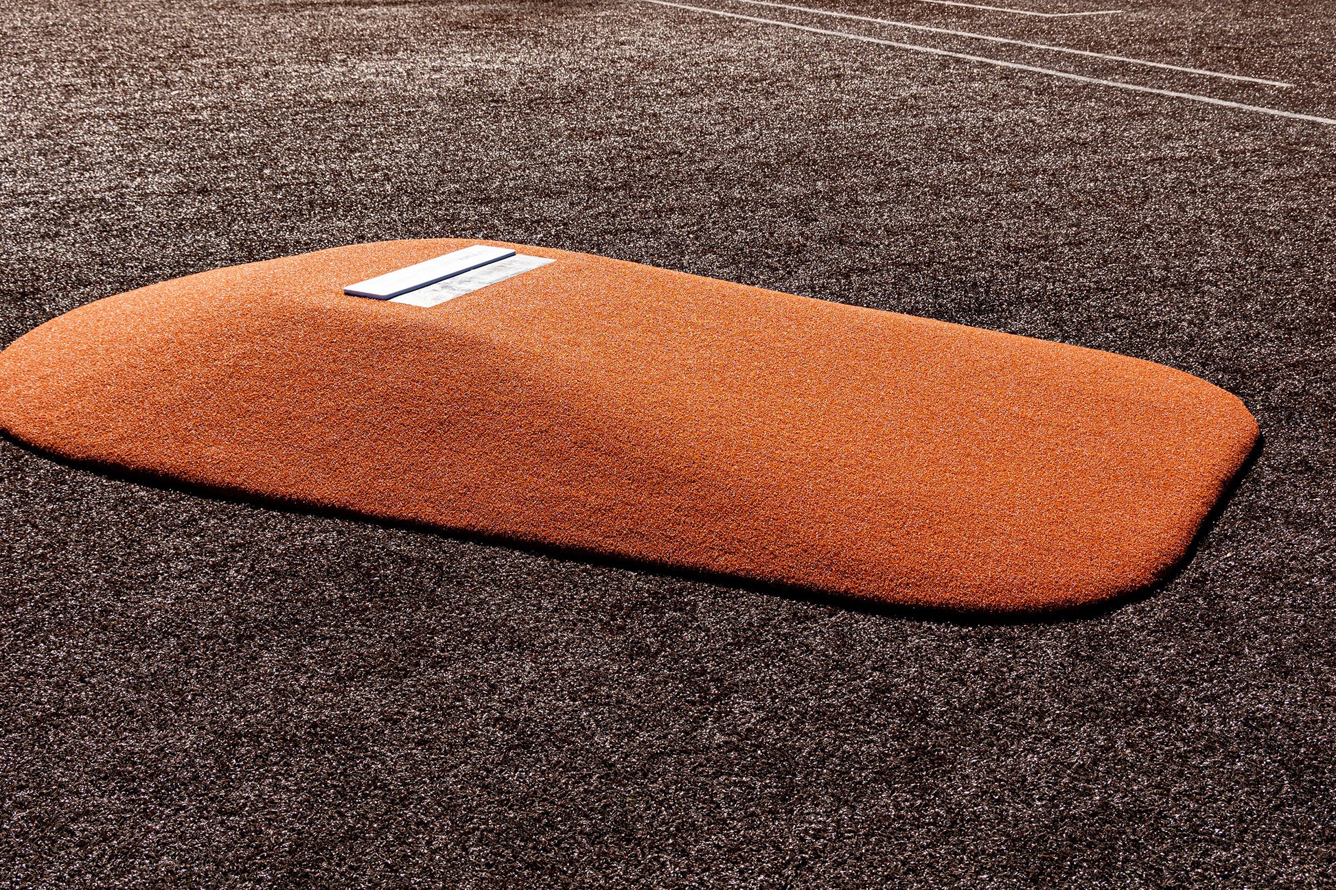 The Portolite 10″ One-Piece Game Mound, a heavy-duty regulation pitching mound featuring an orange design with a white rubber plate, is positioned on a brown artificial turf baseball field; visible white foul lines in the background make it ideal for high school baseball games.