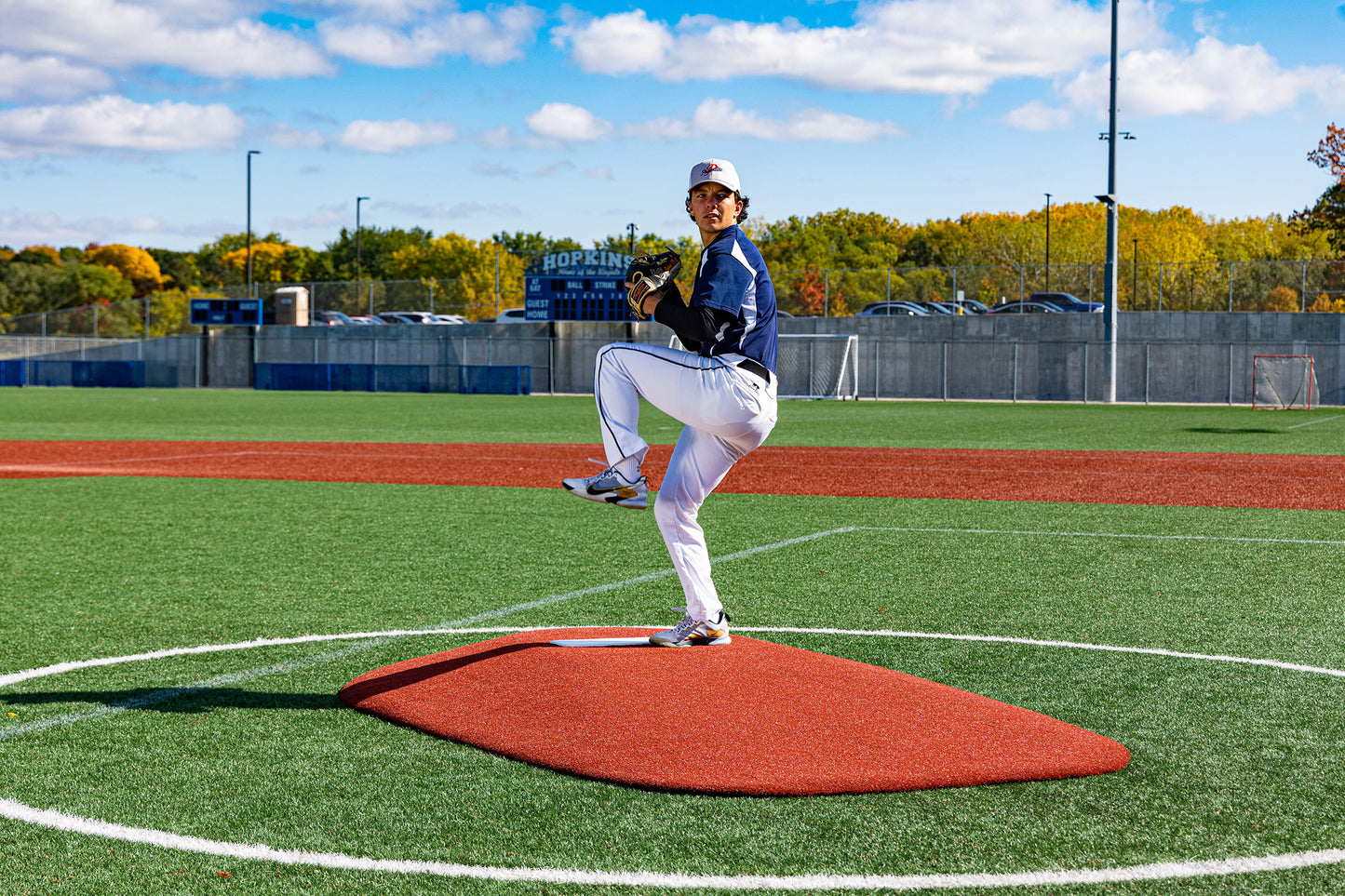 A high school baseball player in uniform stands on a Portolite 10″ One-Piece Game Mound, mid-pitch, on a green field with a scoreboard, cars, and autumn trees visible in the background beneath a partly cloudy sky.