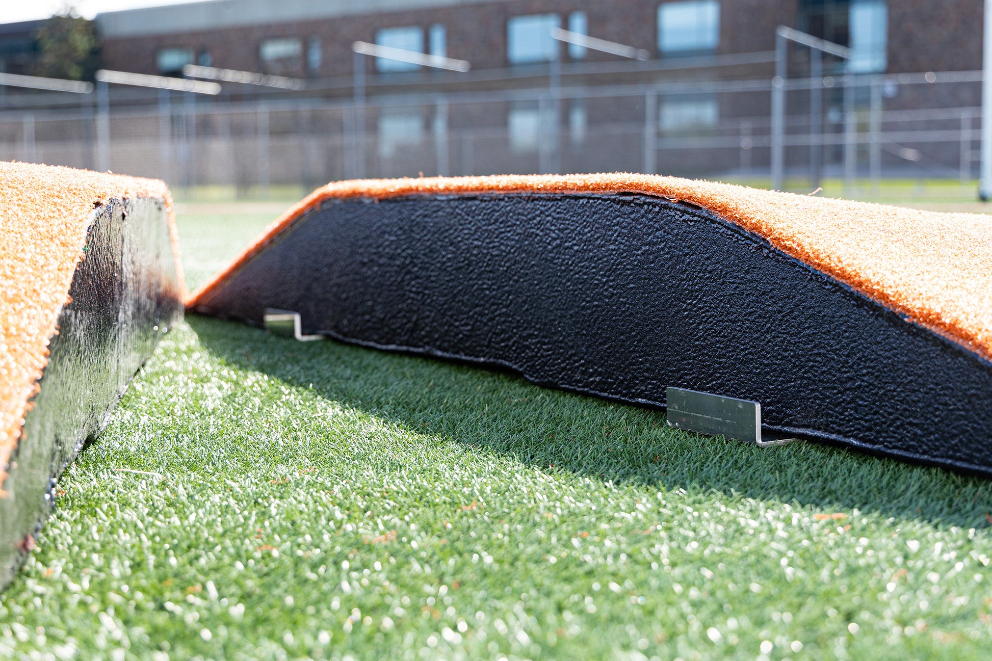 Two Portolite 10″ Two-Piece Game Mounds | Portable Pitching Mounds, featuring black surfaces with bright orange edges, rest on artificial turf that mimics an AstroTurf mound. The photo, taken at ground level, highlights the detailed textures of both the pitching mounds and the synthetic grass, while a building and fences appear blurred in the background.