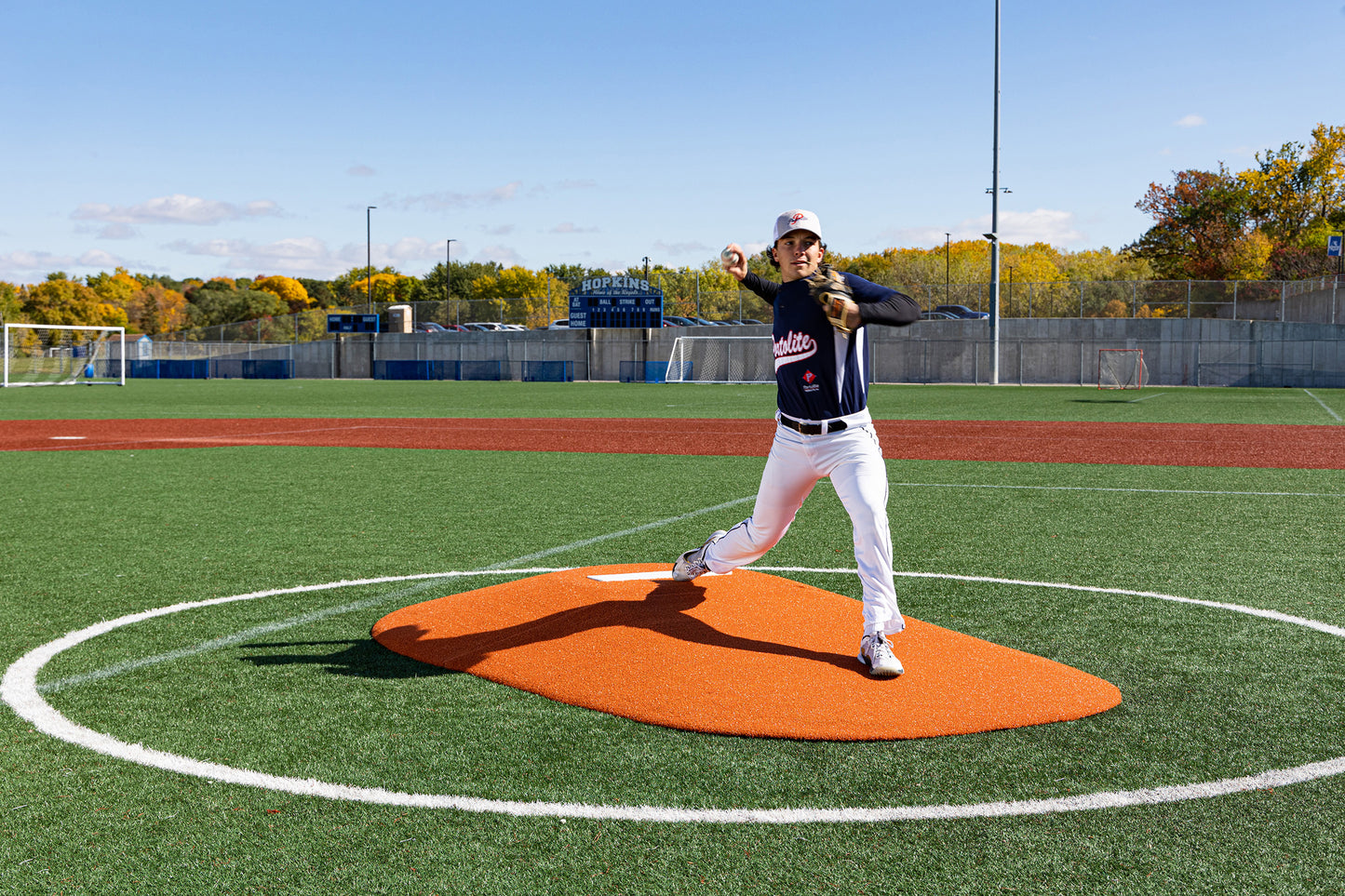 A baseball player in a dark jersey and white pants is pitching from the Portolite 10″ Two-Piece Game Mound | Portable Pitching Mounds, which is set up on an orange mound at the center of a green baseball field, with trees and a scoreboard visible in the background.