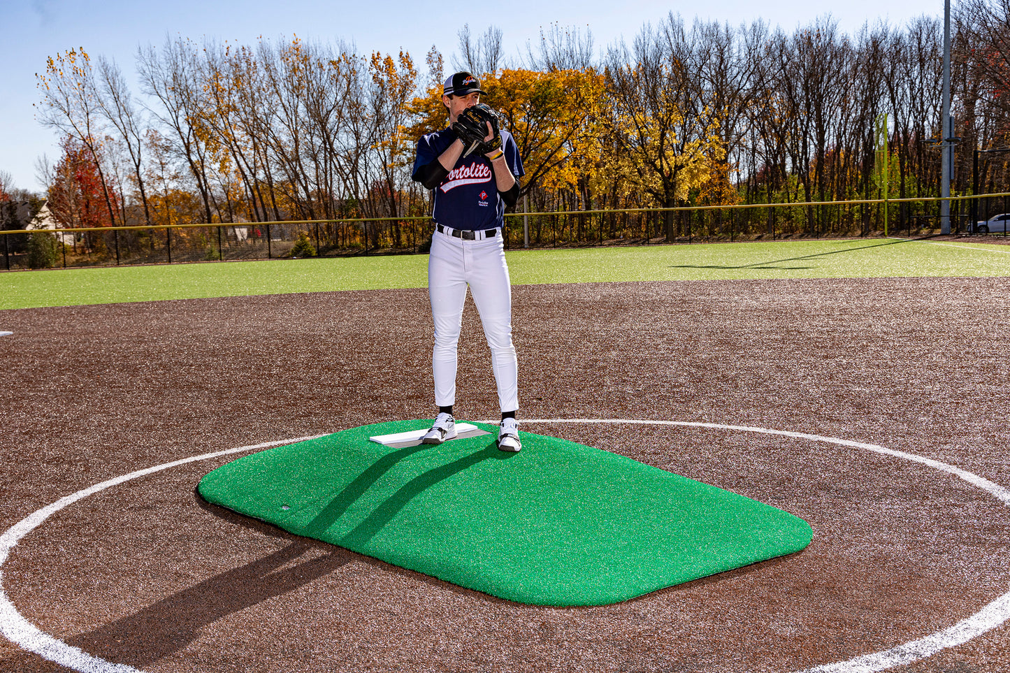 A baseball player in uniform stands on a Portolite 10″ Two-Piece Game Mound | Portable Pitching Mounds—a green portable pitching mound—holding a glove to his face on a sunny day, with autumn trees in the background.
