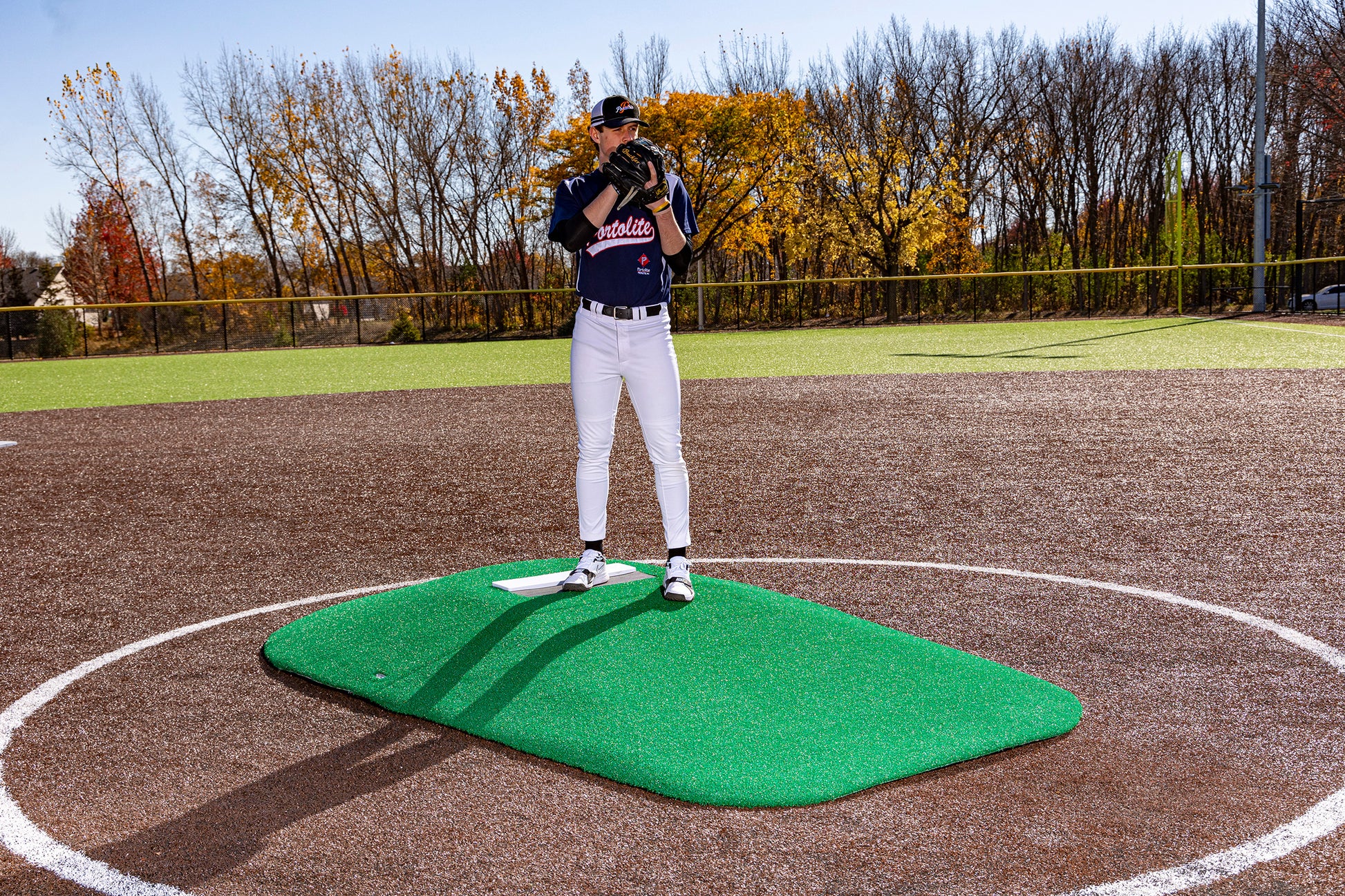 A baseball player in uniform stands on a Portolite 10″ Two-Piece Game Mound | Portable Pitching Mounds—a green portable pitching mound—holding a glove to his face on a sunny day, with autumn trees in the background.