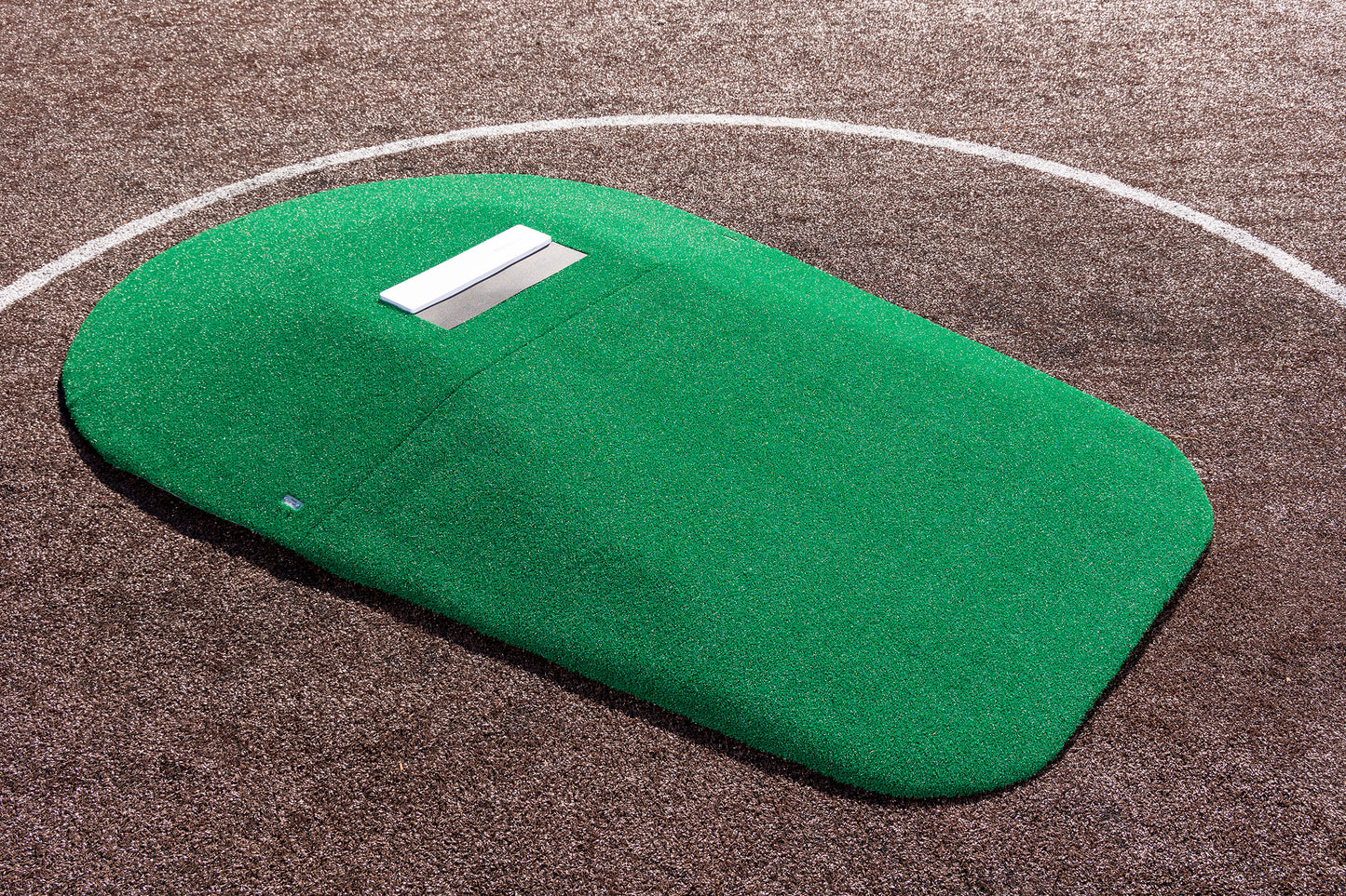 A Portolite 10″ Two-Piece Game Mound portable pitching mound with green AstroTurf and a white rubber plate is positioned on a brown baseball or softball field, within a white chalk pitching circle.