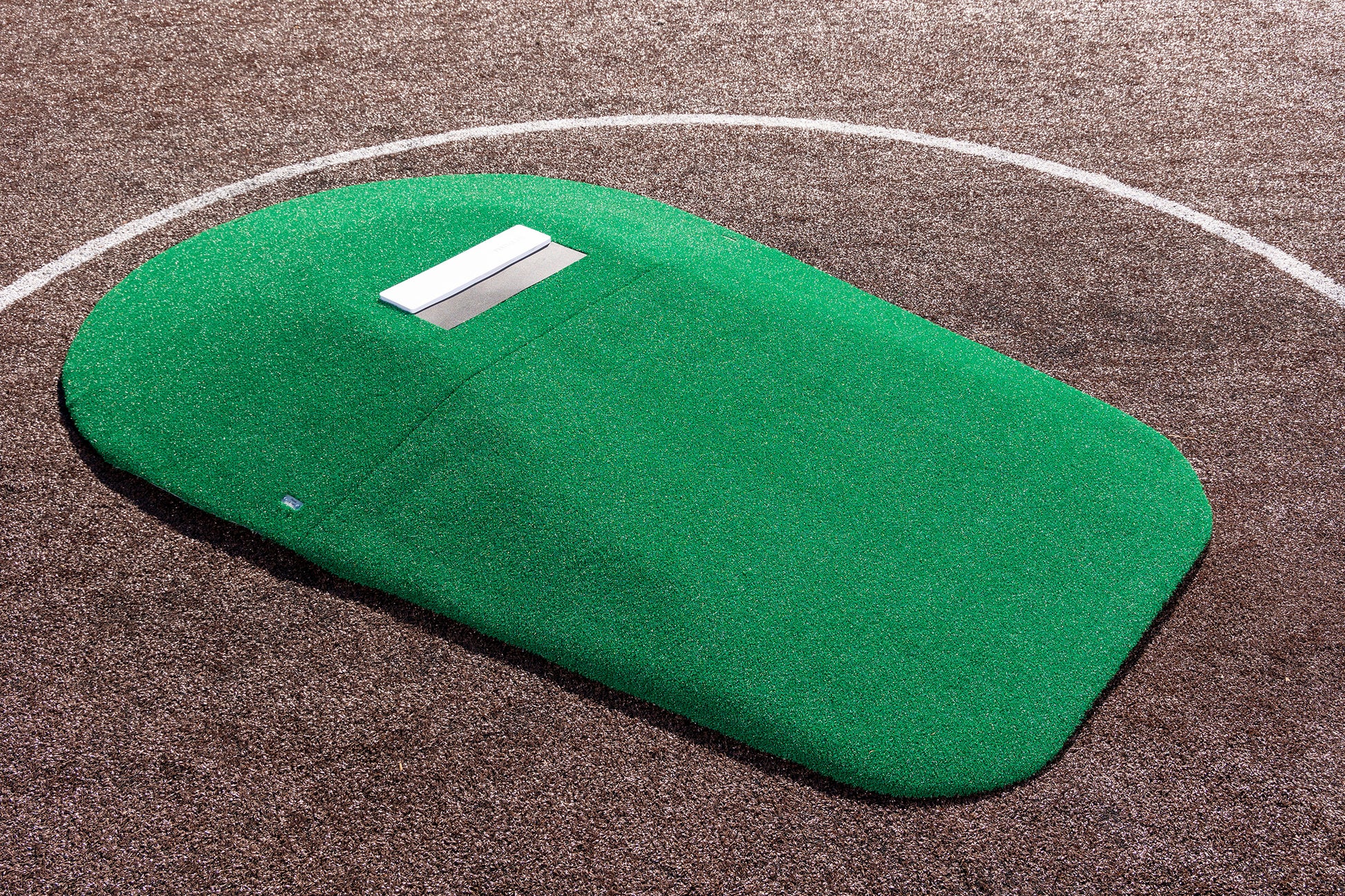 A Portolite 10″ Two-Piece Game Mound portable pitching mound with green AstroTurf and a white rubber plate is positioned on a brown baseball or softball field, within a white chalk pitching circle.
