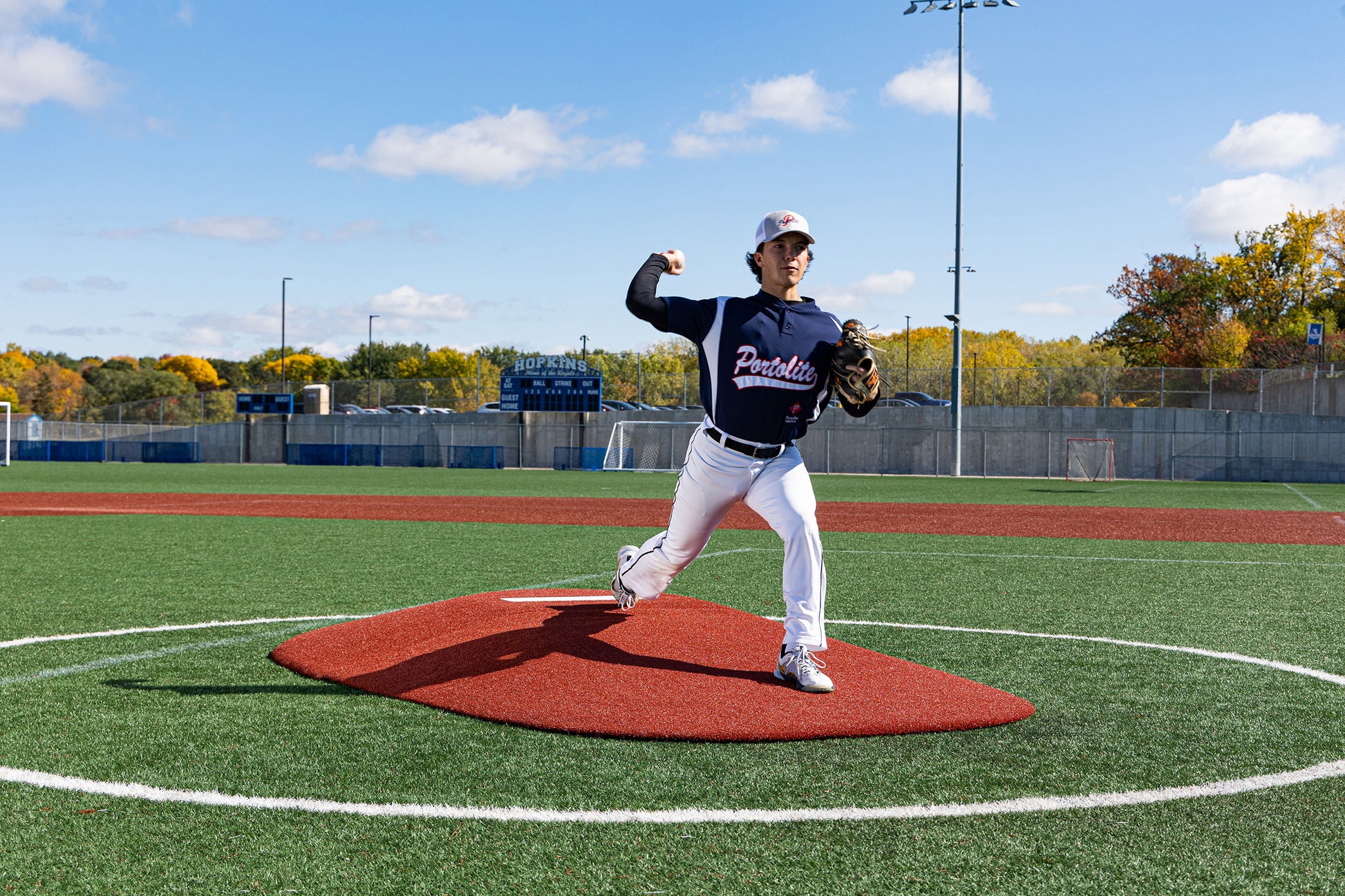 A baseball player in a dark jersey and white pants is pitching on the Portolite 10″ Two-Piece Game Mound | Portable Pitching Mounds at an outdoor field beneath a blue sky with autumn trees in the background.