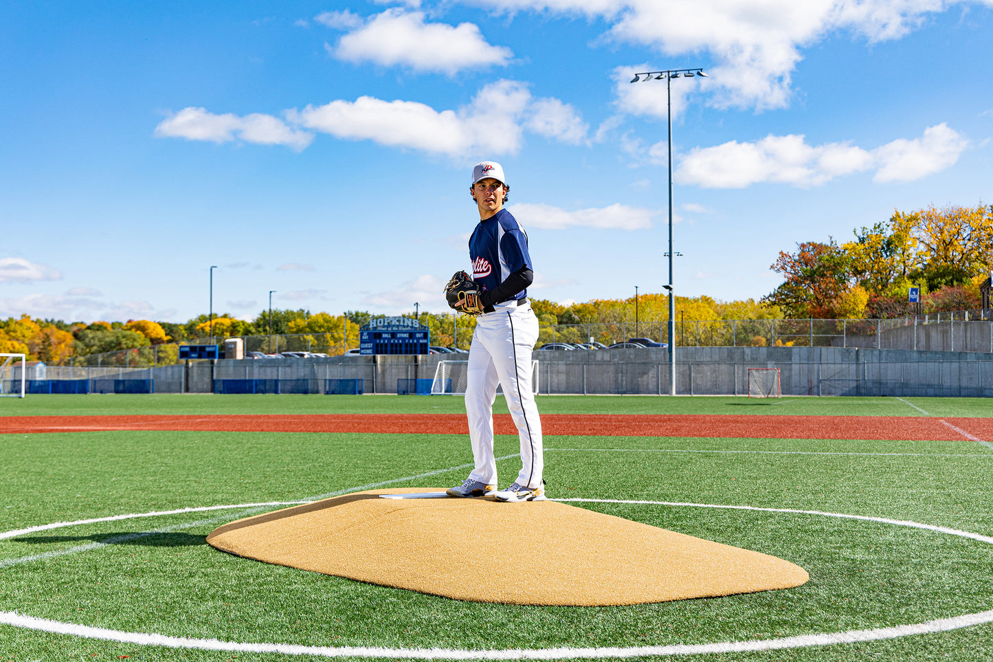 A baseball player stands on a Portolite 10″ Two-Piece Game Mound | Portable Pitching Mounds on an outdoor field beneath a bright blue sky with scattered clouds and autumn trees in the background.