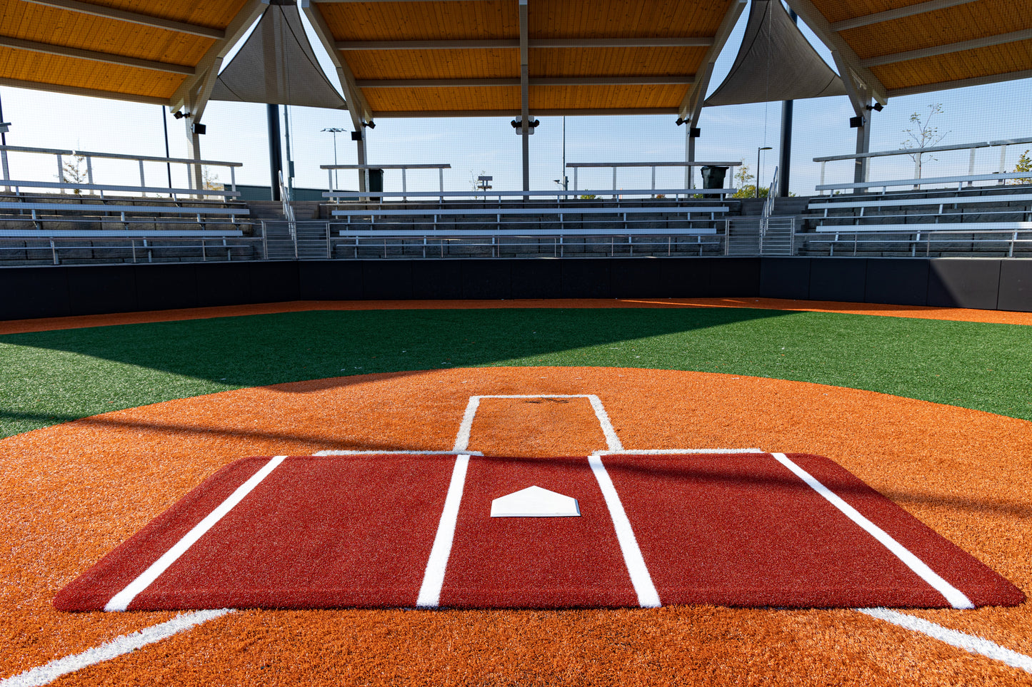 The home plate area of an empty baseball field with artificial turf showcases the Portolite Full Batter’s Mat | 12′ x 6′ No Slip Batter’s Mat featuring SpikeTek technology, under a shaded roof and facing vacant bleachers on a sunny day—an example of advanced training facilities at their best.