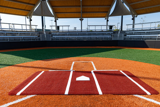 The home plate area of an empty baseball field with artificial turf showcases the Portolite Full Batter’s Mat | 12′ x 6′ No Slip Batter’s Mat featuring SpikeTek technology, under a shaded roof and facing vacant bleachers on a sunny day—an example of advanced training facilities at their best.