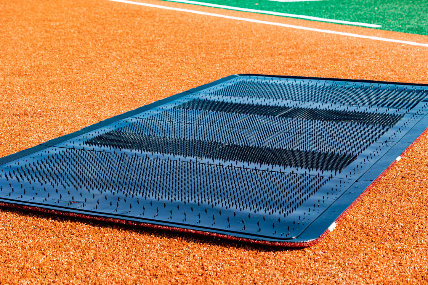 A close-up of the Portolite Full Batter’s Mat | 12′ x 6′ No Slip Batter’s Mat is shown on a baseball or softball field’s infield dirt, illustrating its use for providing a stable, slip-resistant surface during training and games. Green turf appears in the background, emphasizing the mat’s quality and suitability for advanced training facilities.