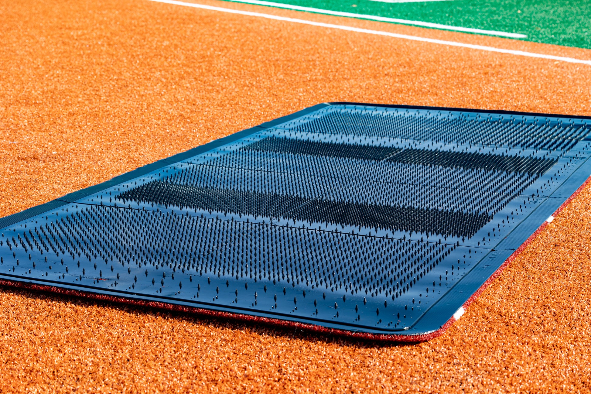 A close-up of the Portolite Full Batter’s Mat | 12′ x 6′ No Slip Batter’s Mat is shown on a baseball or softball field’s infield dirt, illustrating its use for providing a stable, slip-resistant surface during training and games. Green turf appears in the background, emphasizing the mat’s quality and suitability for advanced training facilities.