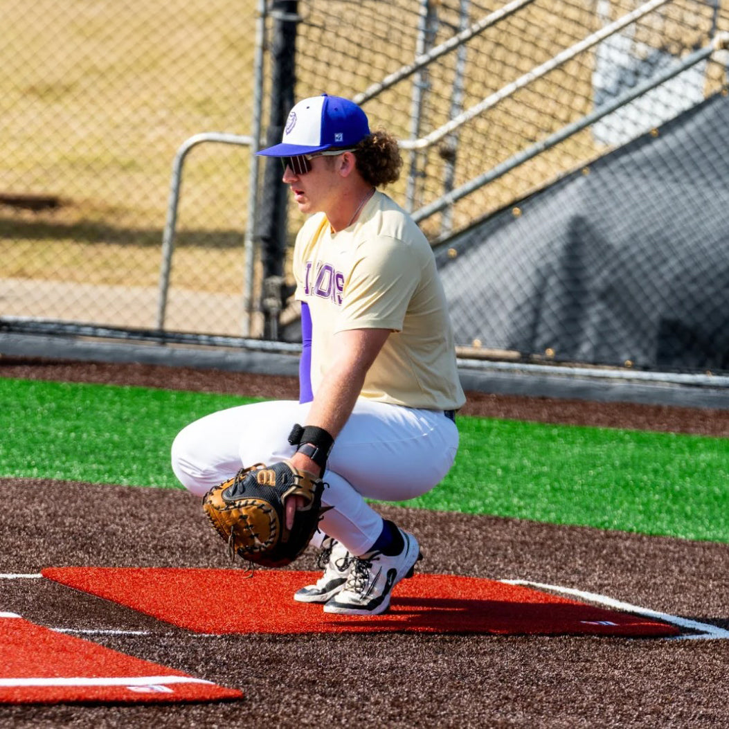 A baseball player in uniform squats with a glove, preparing to catch, on a field near home plate where the Portolite Catcher’s Mat | SpikeTek Turf & Dirt Protection Mat is placed on the red and brown turf. A fence and stairs can be seen in the background.